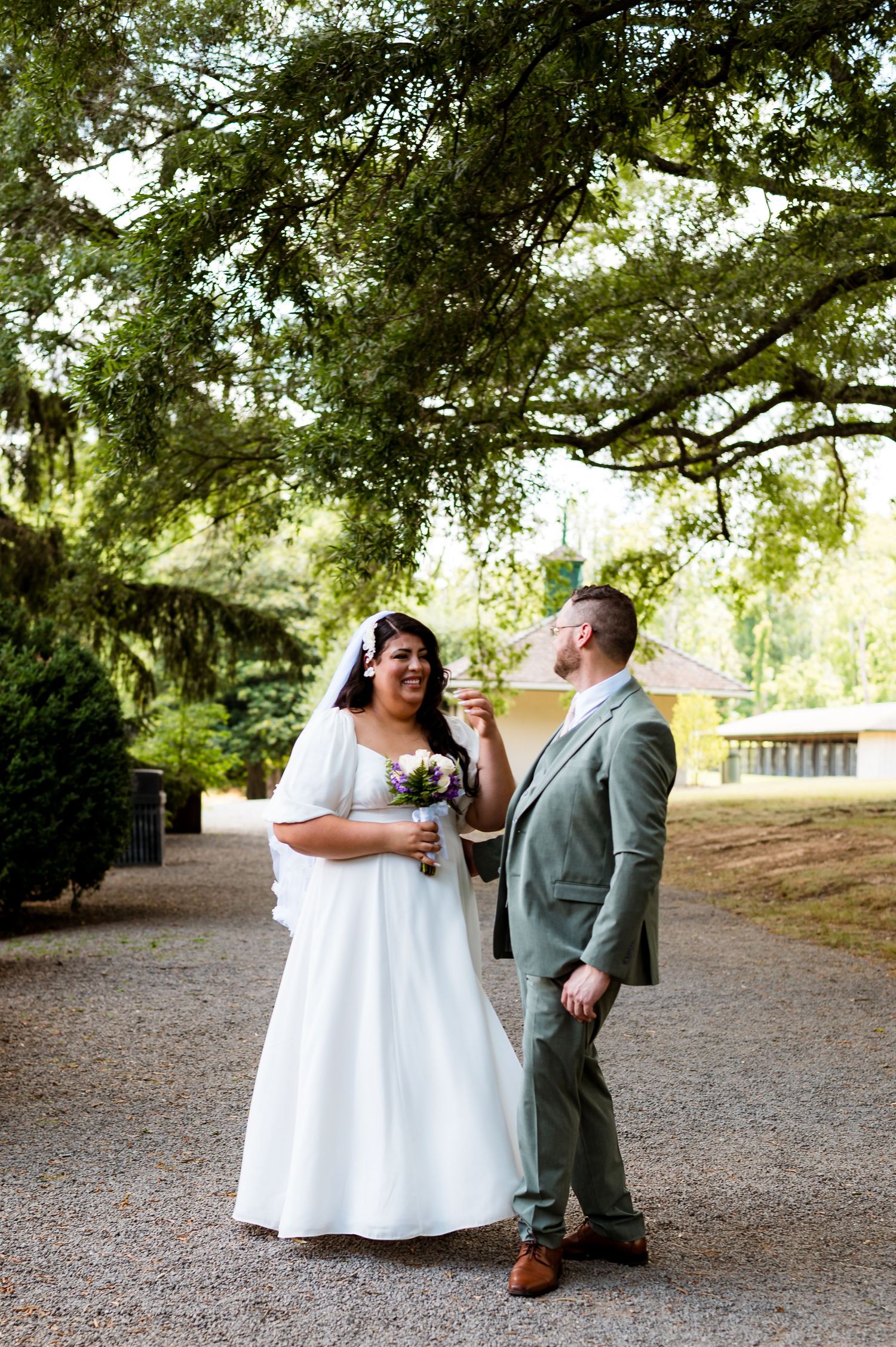 A couple in wedding attire shares a joyful moment under a large tree on a gravel path.