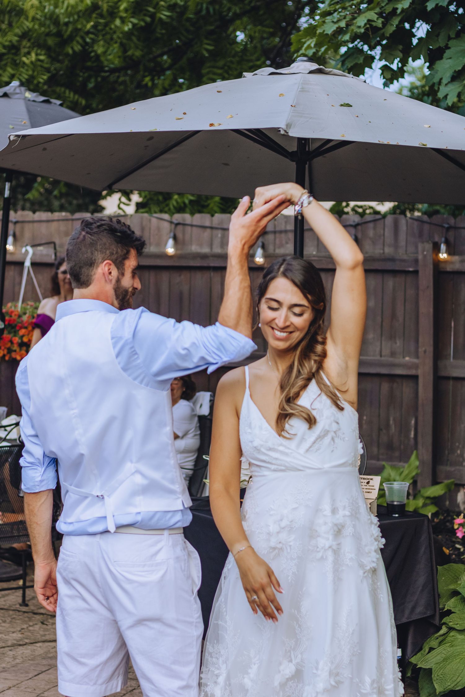 Couple wearing light colored outfits dance together in a garden setting, twirling and smiling.