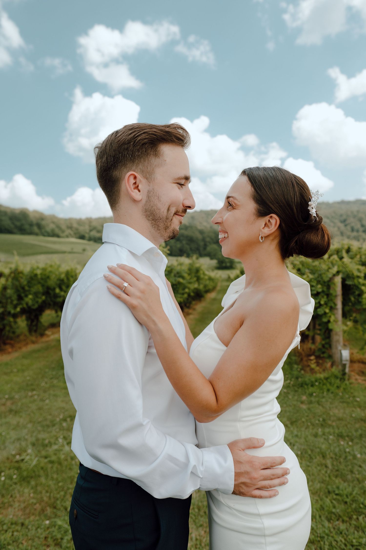 A joyful couple embraces outdoors at a scenic vineyard on their wedding day against a blue summer sky.