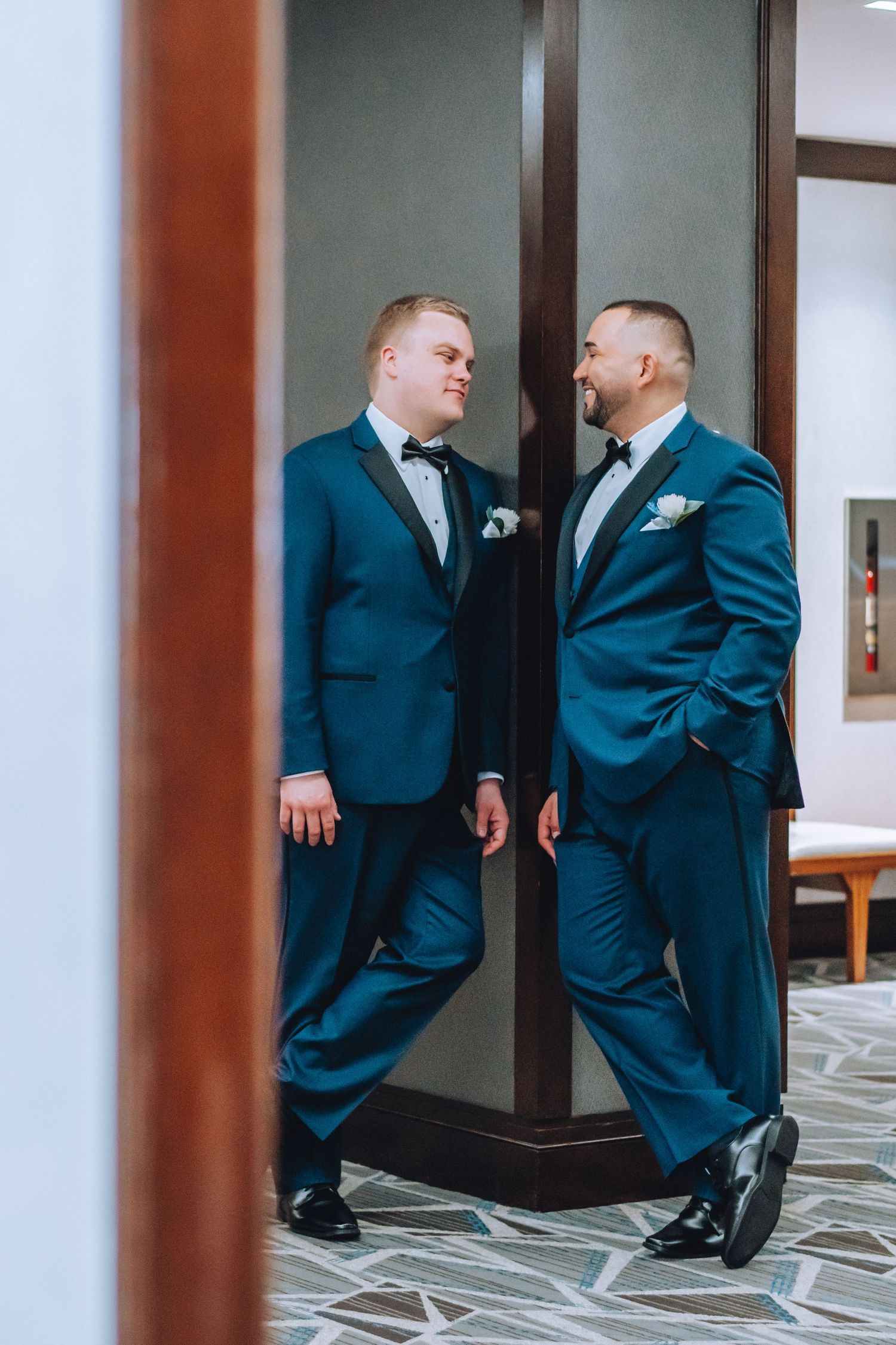 Two grooms in matching navy blue suits share an intimate moment together in a quiet hotel hallway.