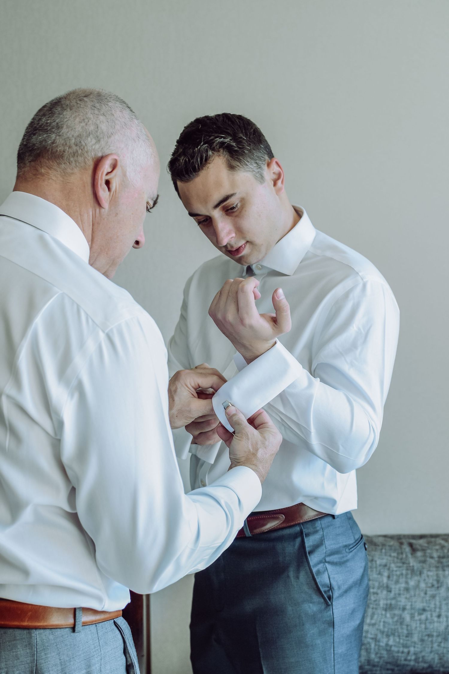 Two men in white dress shirts preparing for formal event, one helping the other adjust cufflinks.