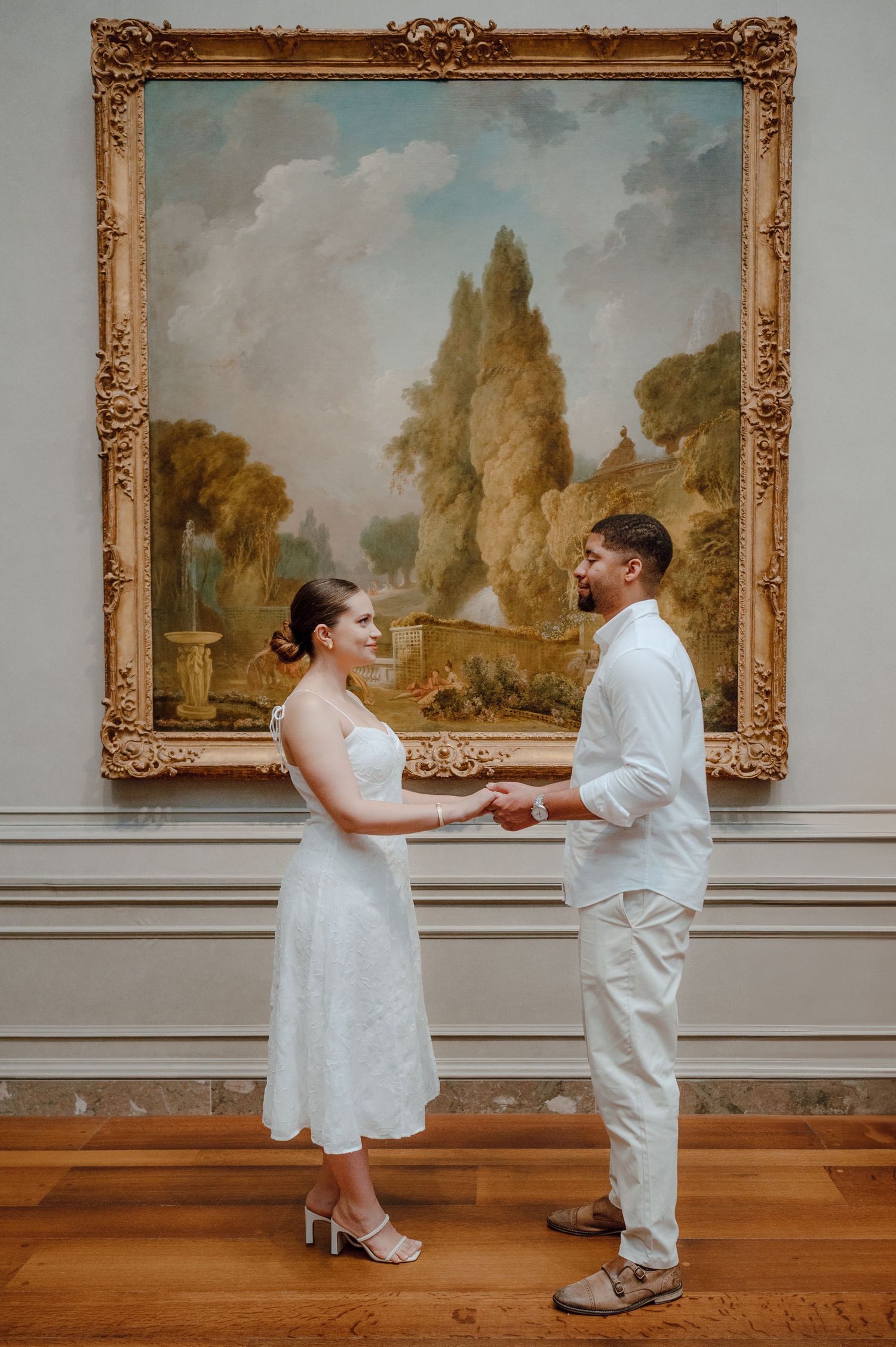 A couple shares a romantic moment in a museum gallery in front of a large pastoral landscape painting in an ornate gold frame.