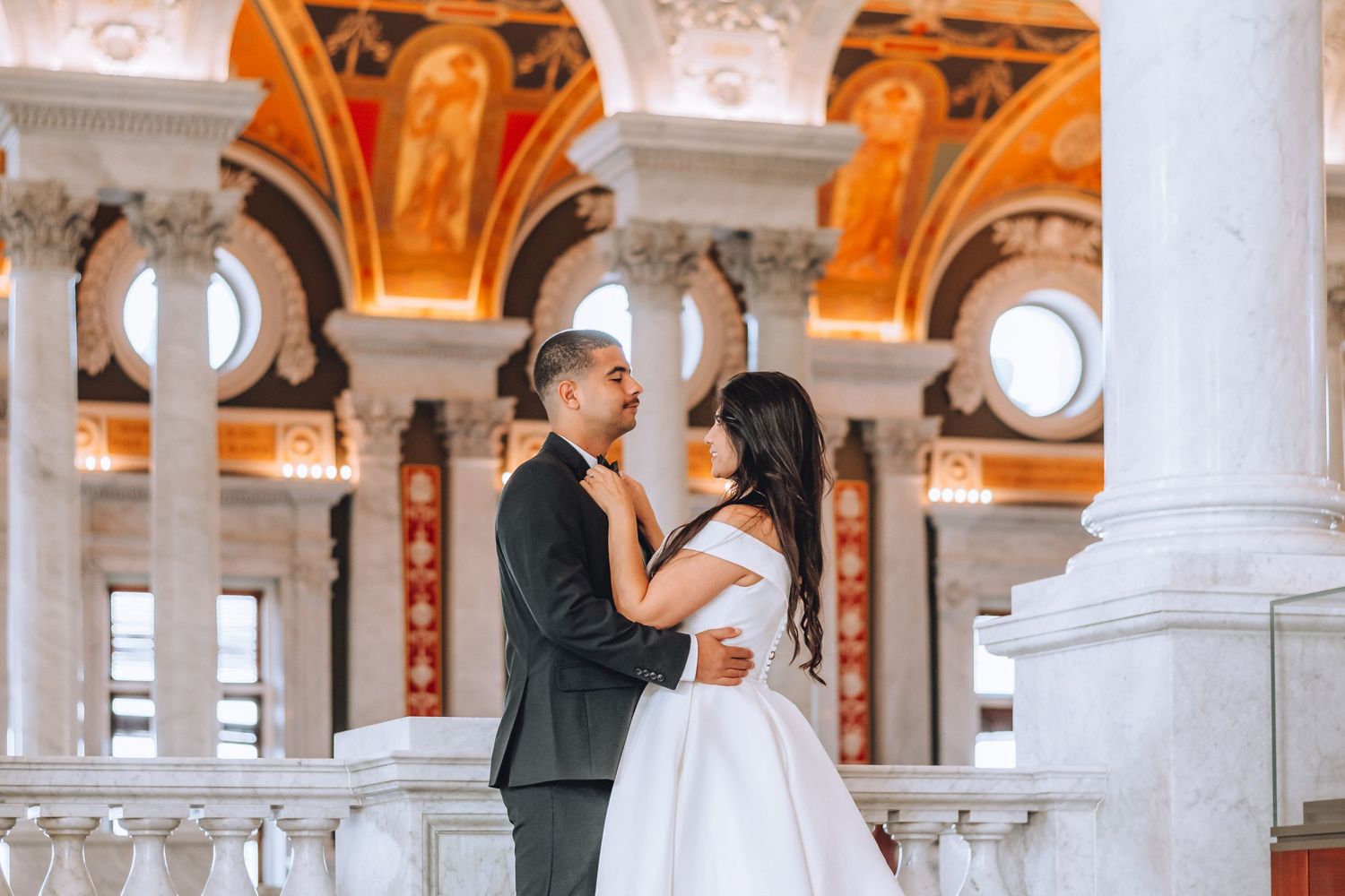 A couple shares a tender moment on an elegant balcony with classical architecture and orange-tinted ceilings.