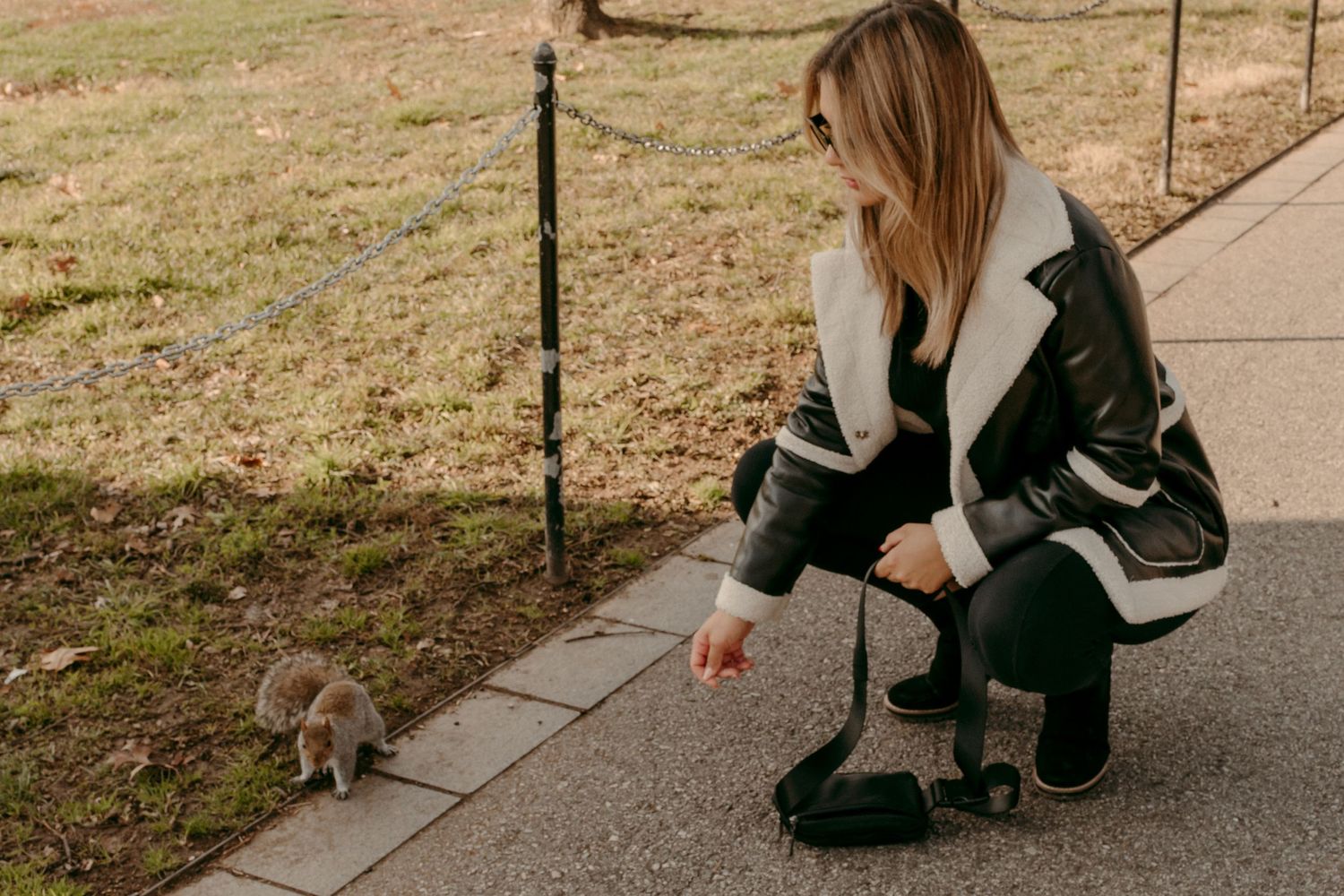Person in leather jacket crouches down to feed a squirrel on a park pathway.