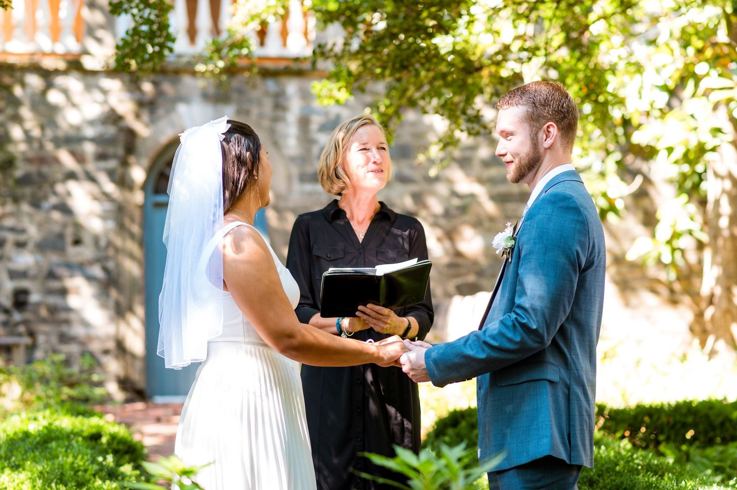 Outdoor wedding ceremony with officiant joining couple in marriage in front of stone building.