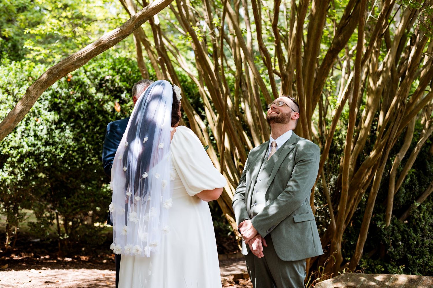 A couple shares a joyful moment during their outdoor garden wedding ceremony beneath a canopy of trees.
