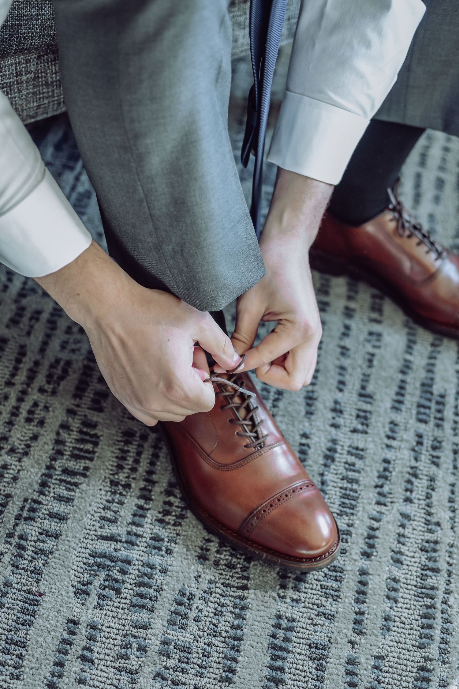 Close up of hands lacing up brown leather dress shoes on a textured carpet.