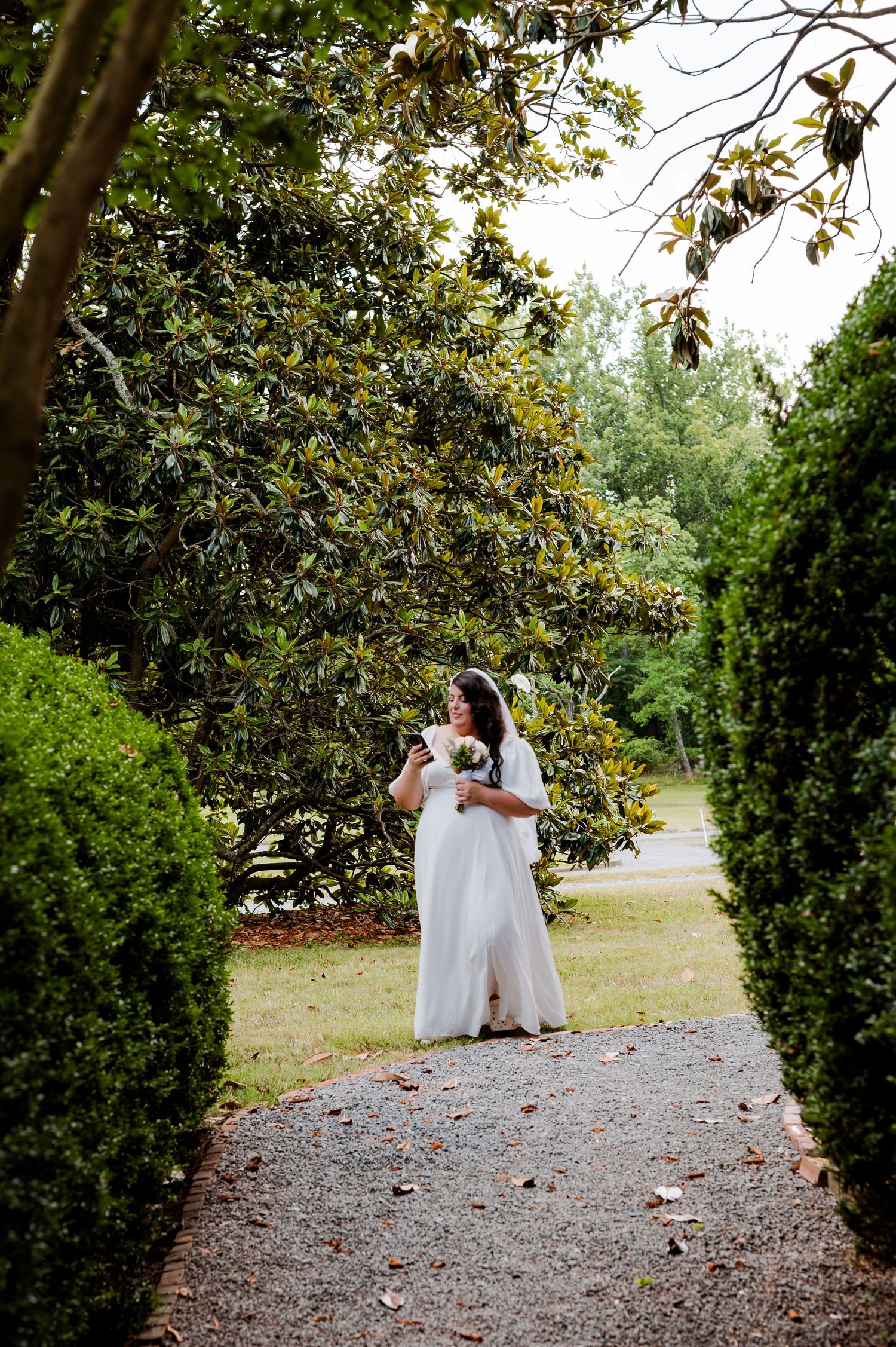 A couple shares a romantic moment on a gravel garden path surrounded by lush green trees and magnolia foliage.