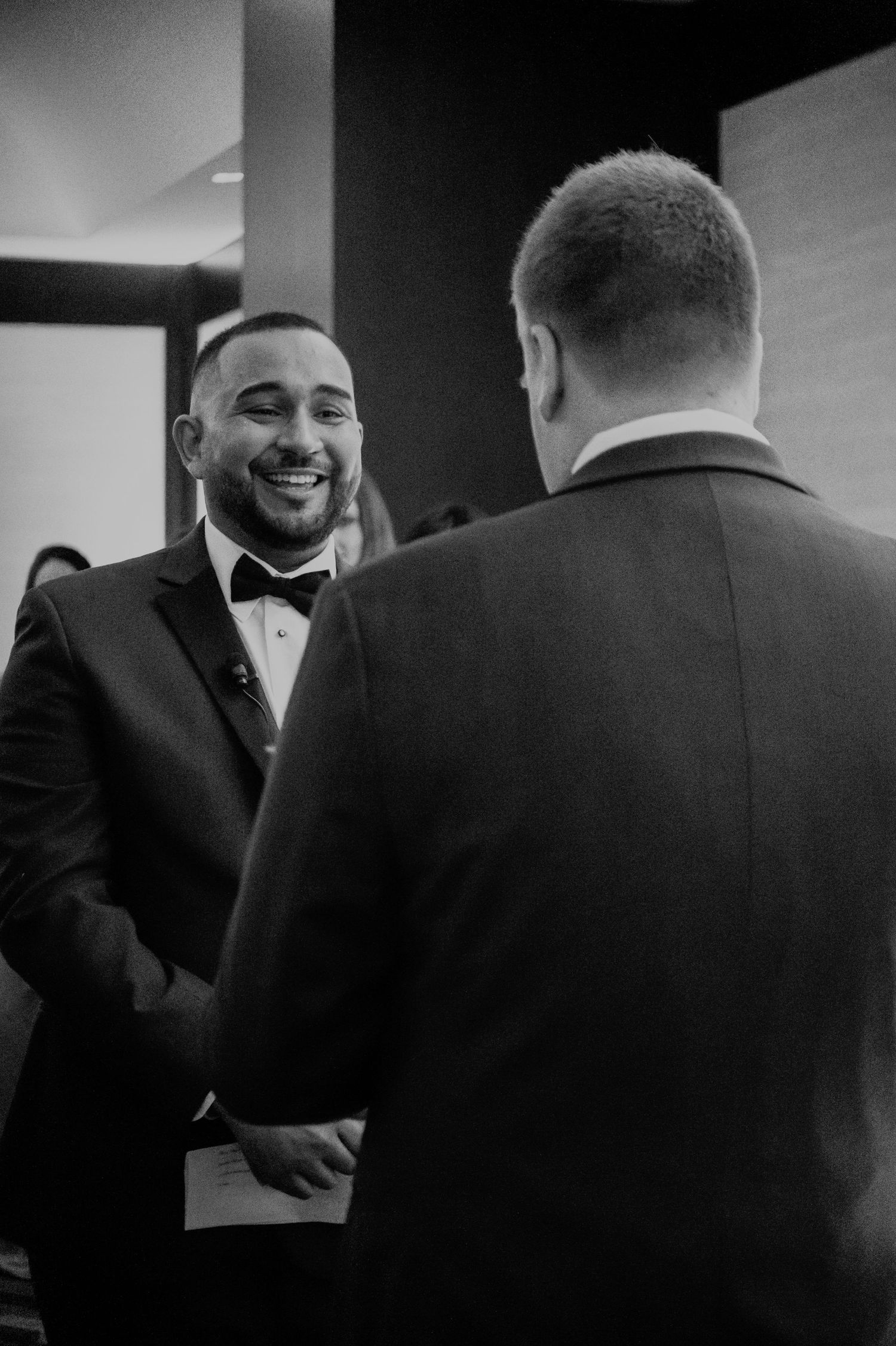 Two men in formal black tuxedos share a conversation and warm smiles at an event with dark background.