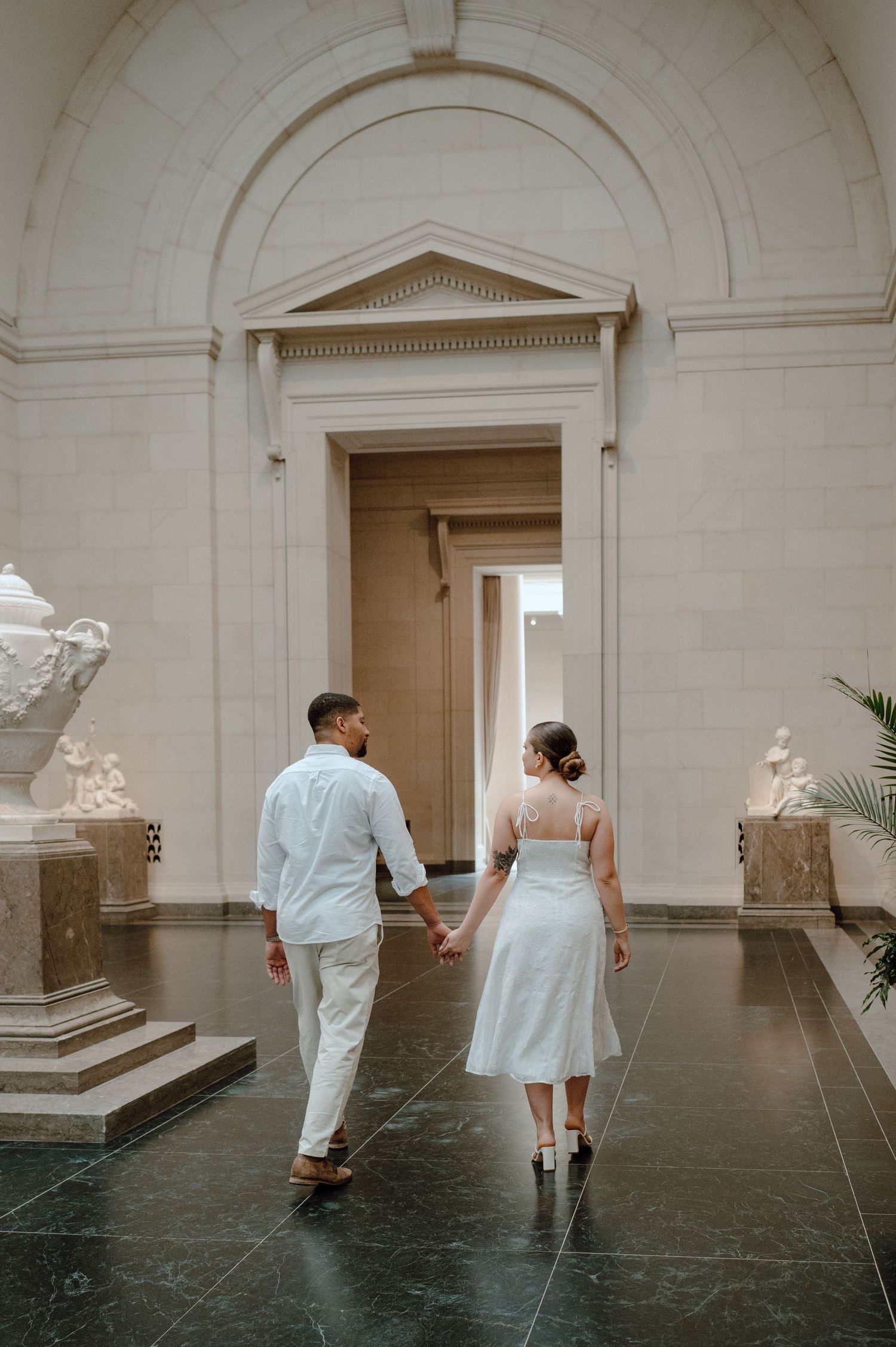 Couple in white outfits walk through elegant museum gallery with marble arches and classical sculptures.