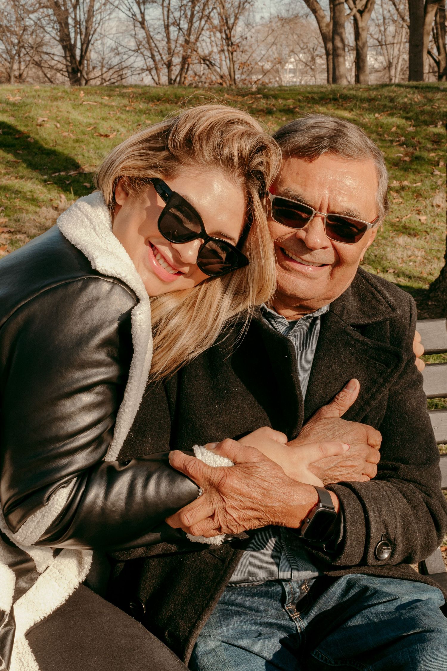 Two people in sunglasses share a warm embrace on a park bench on a sunny day.