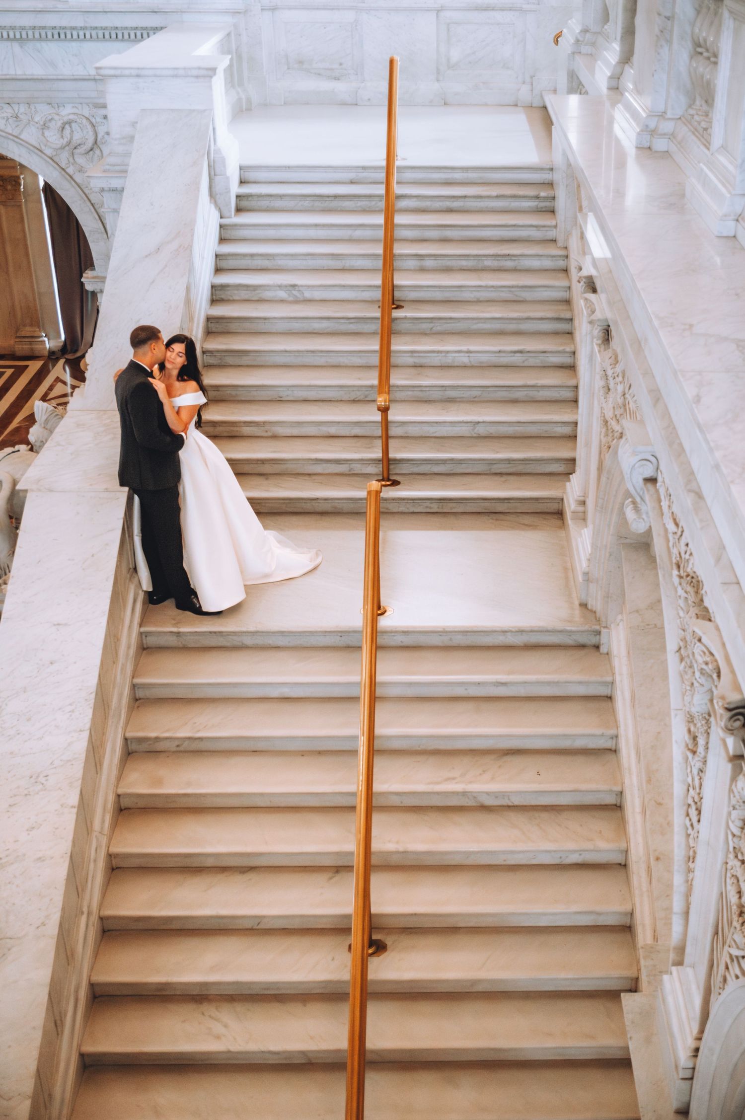 A wedding couple embraces at the bottom of an elegant marble staircase with classical architecture.
