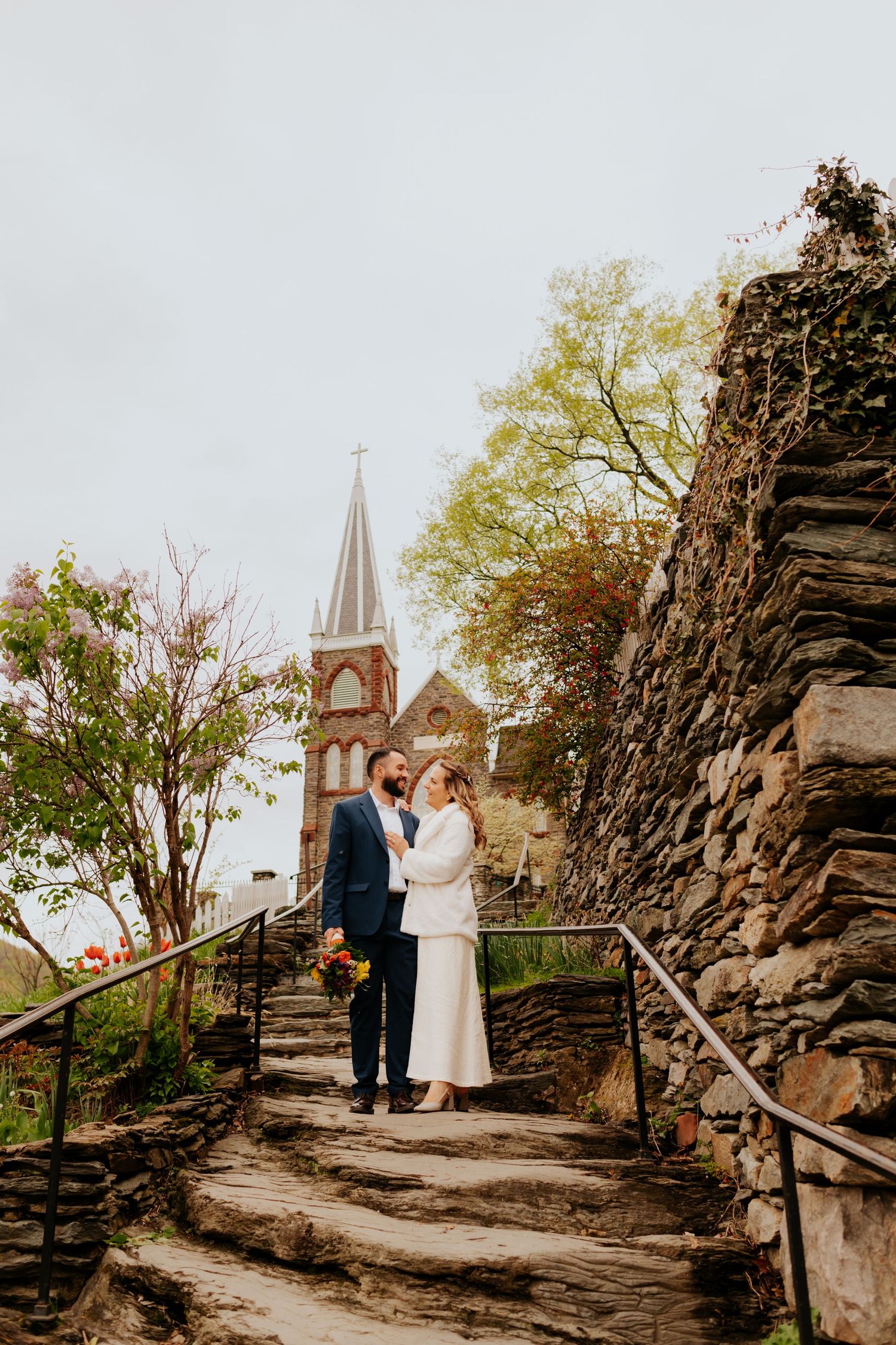 A couple poses on stone steps leading to a historic church with a tall spire in the background.