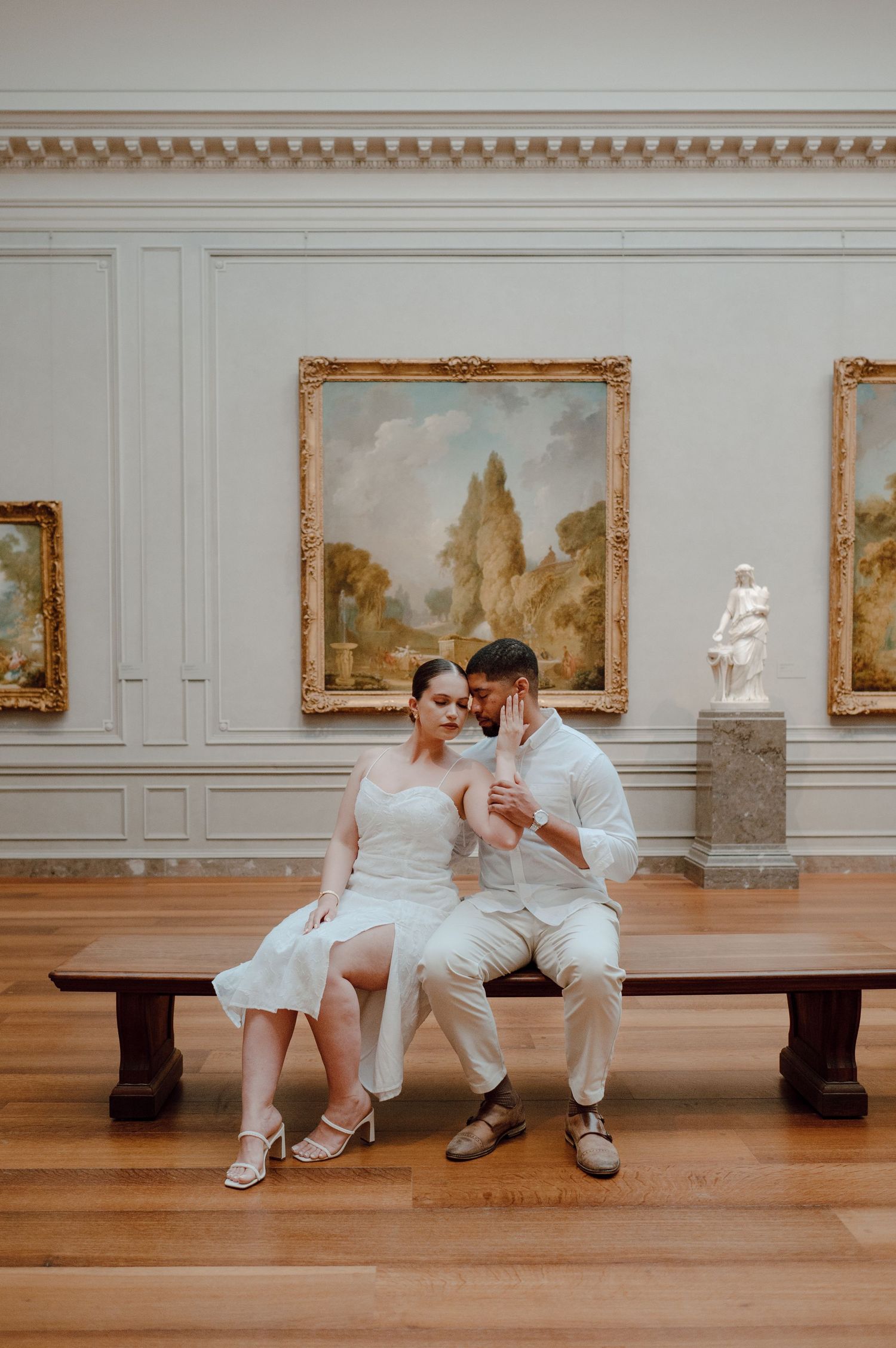 A romantic couple in white outfits sits and interacts on a wooden bench in an art gallery displaying landscape paintings.