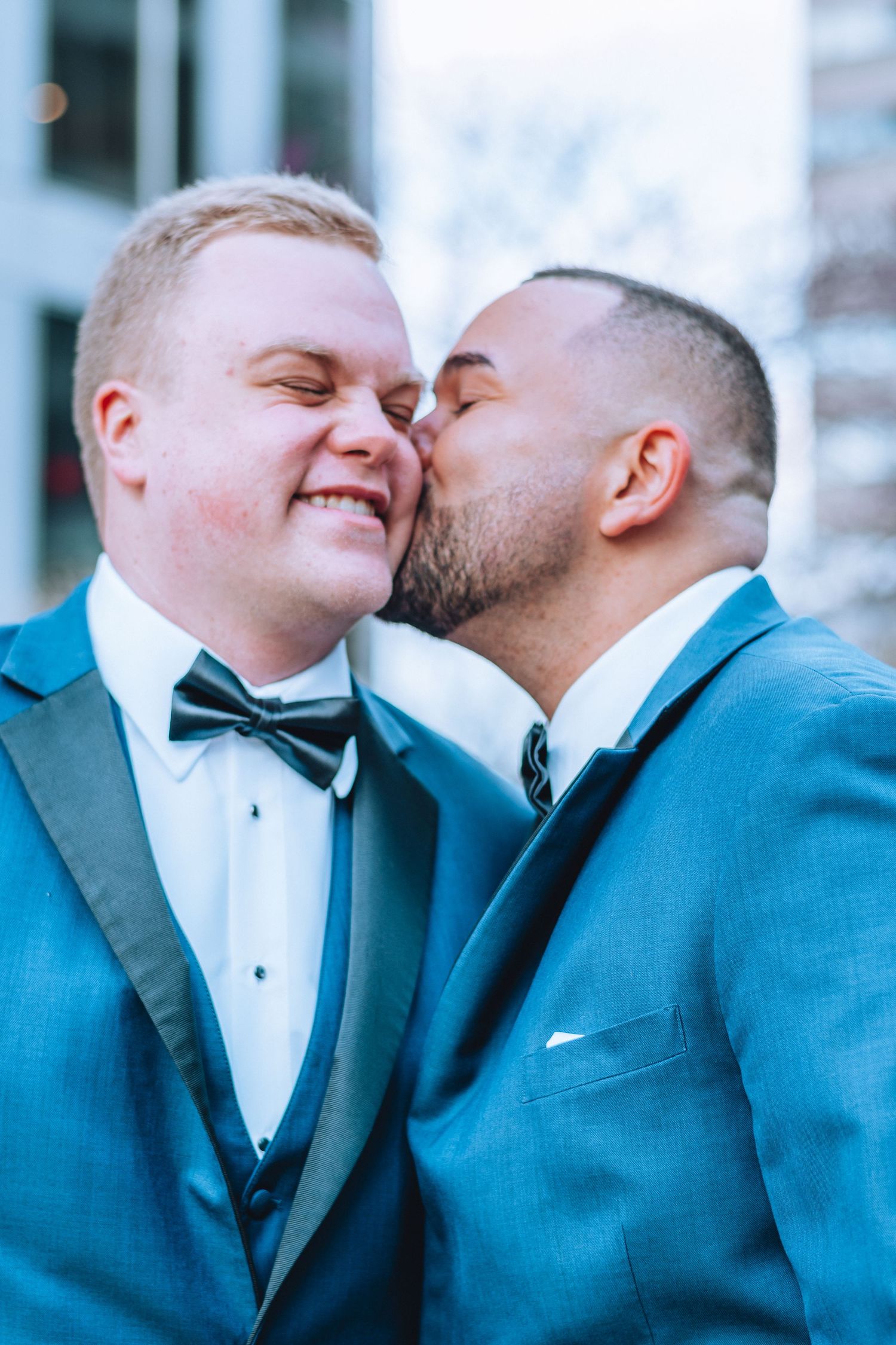 A romantic wedding photo shows two grooms in blue tuxedos sharing a tender moment together outside.