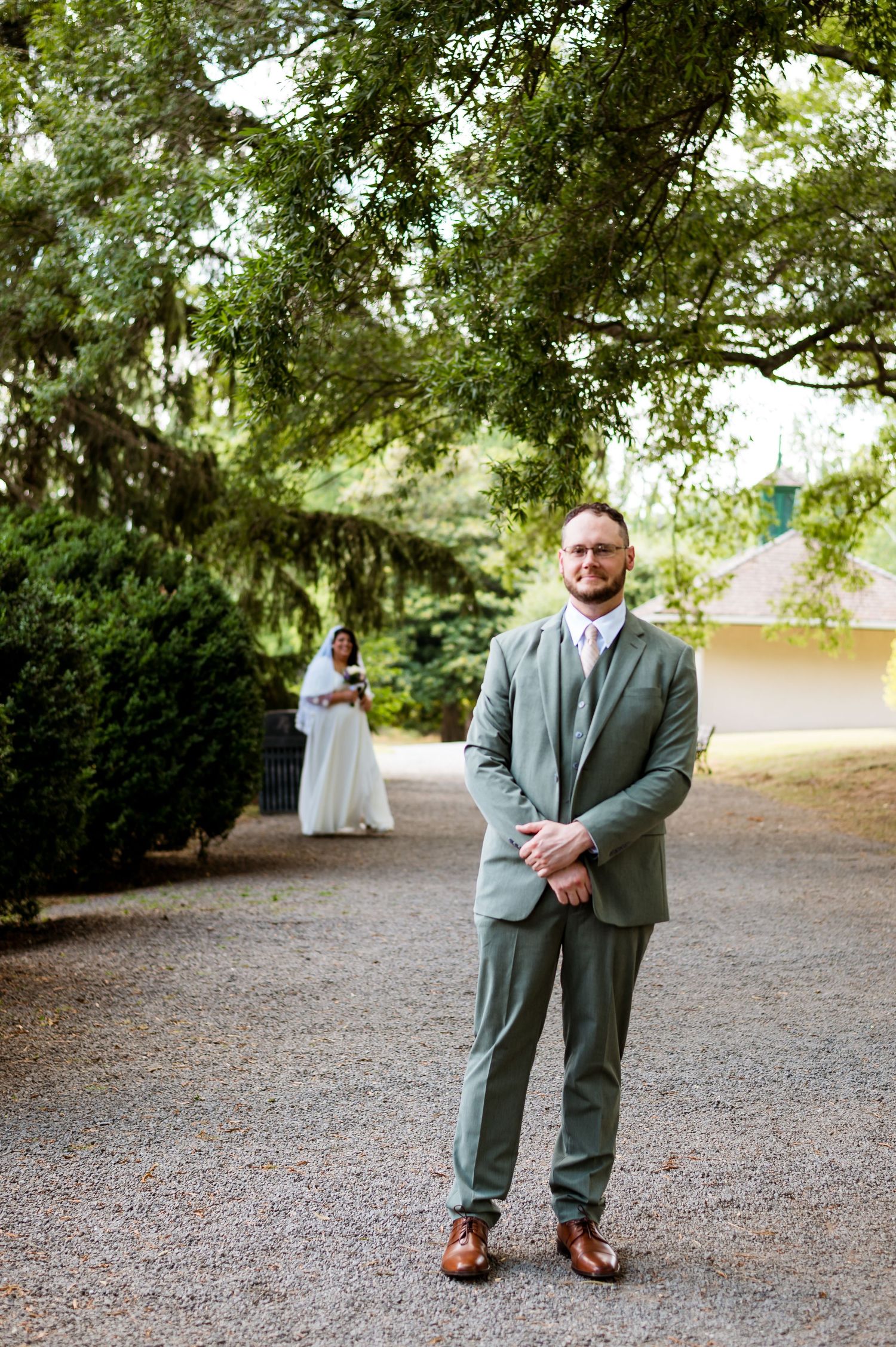 Someone wearing a sage green suit stands on a gravel path with trees in the background.
