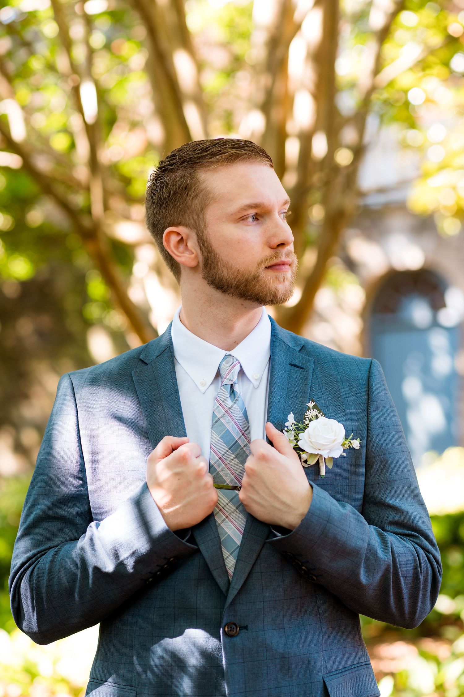 Groom in blue suit and plaid tie with boutonniere poses in natural setting.