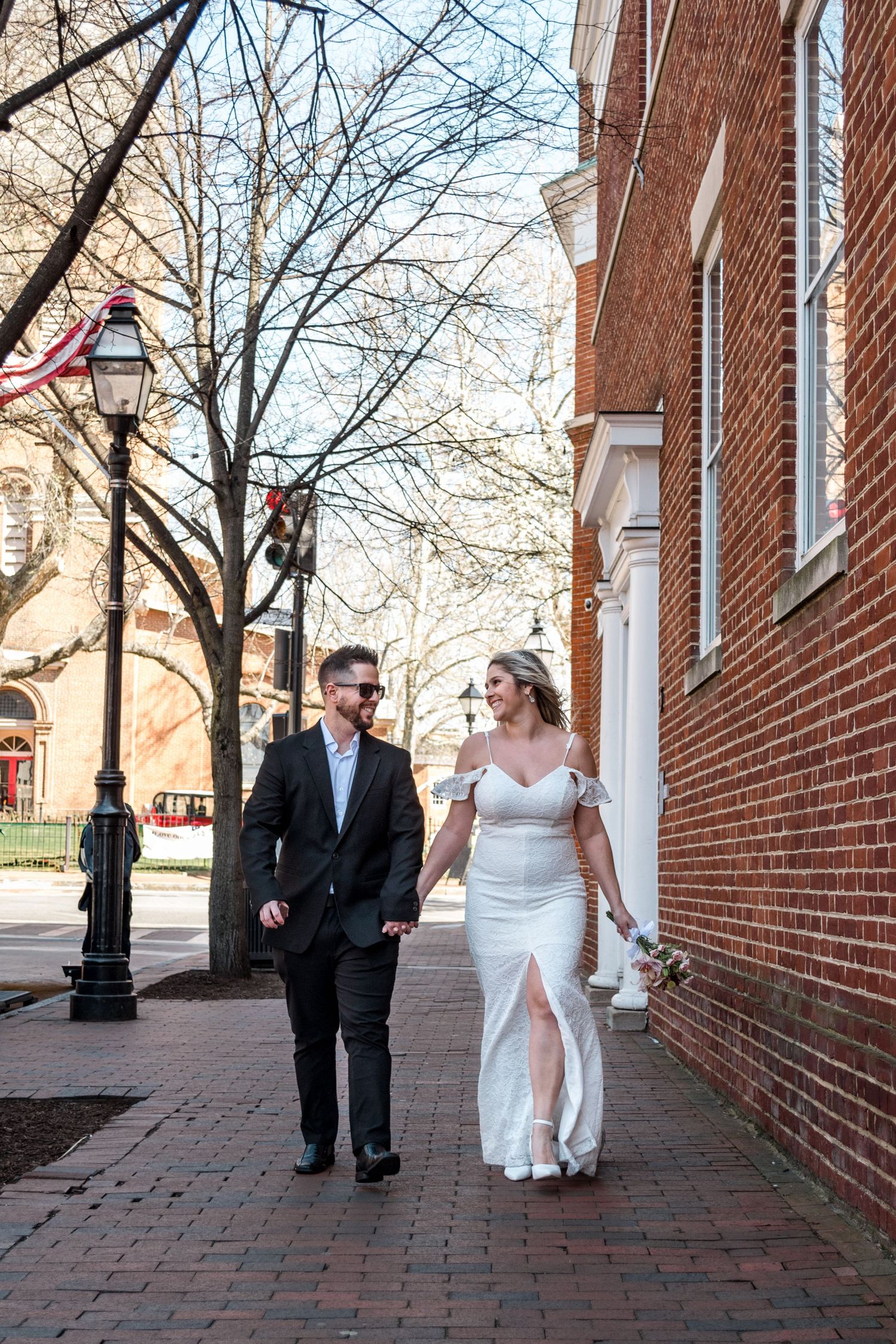 Wedding couple walking together on brick sidewalk beside historic brick building with bare trees.
