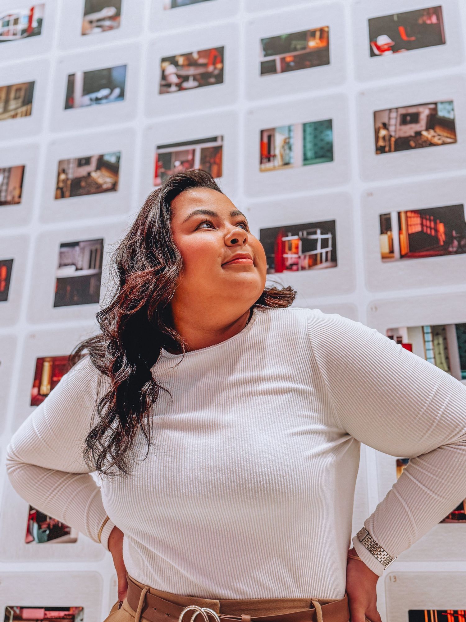 A person in a white ribbed sweater poses against a wall decorated with colorful photo prints.