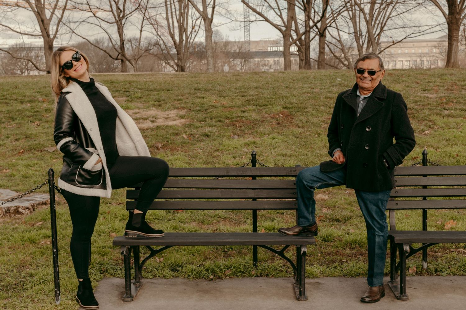 Two people in winter coats posing playfully on opposite ends of a park bench.