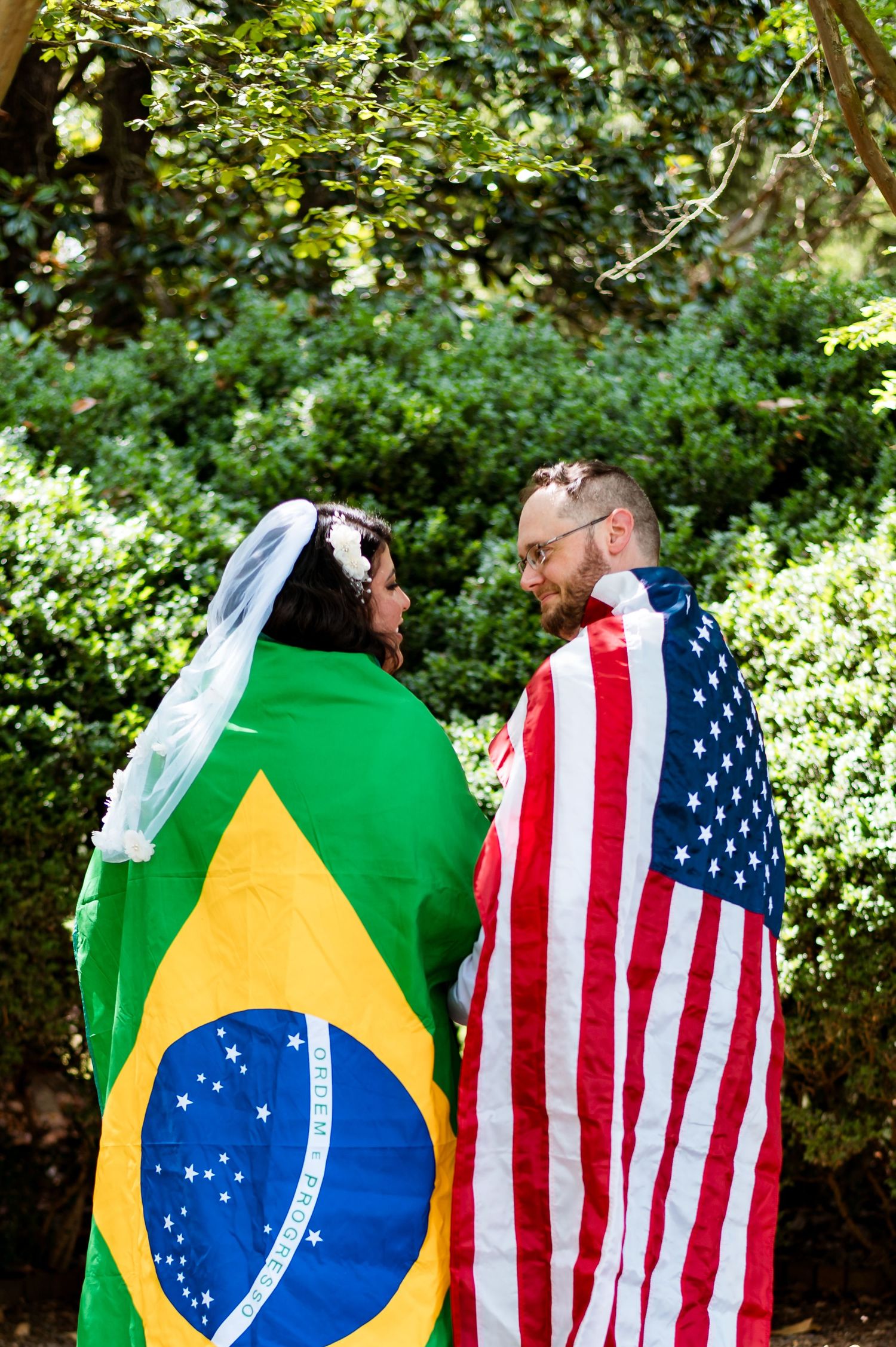 A couple stands wrapped in Brazilian and American flags in a sunlit garden setting.