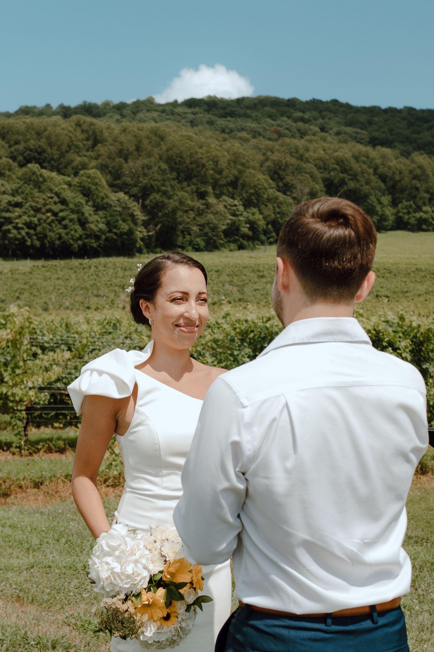 A couple stands together outdoors against a backdrop of green hills with the bride holding a white and peach bouquet.