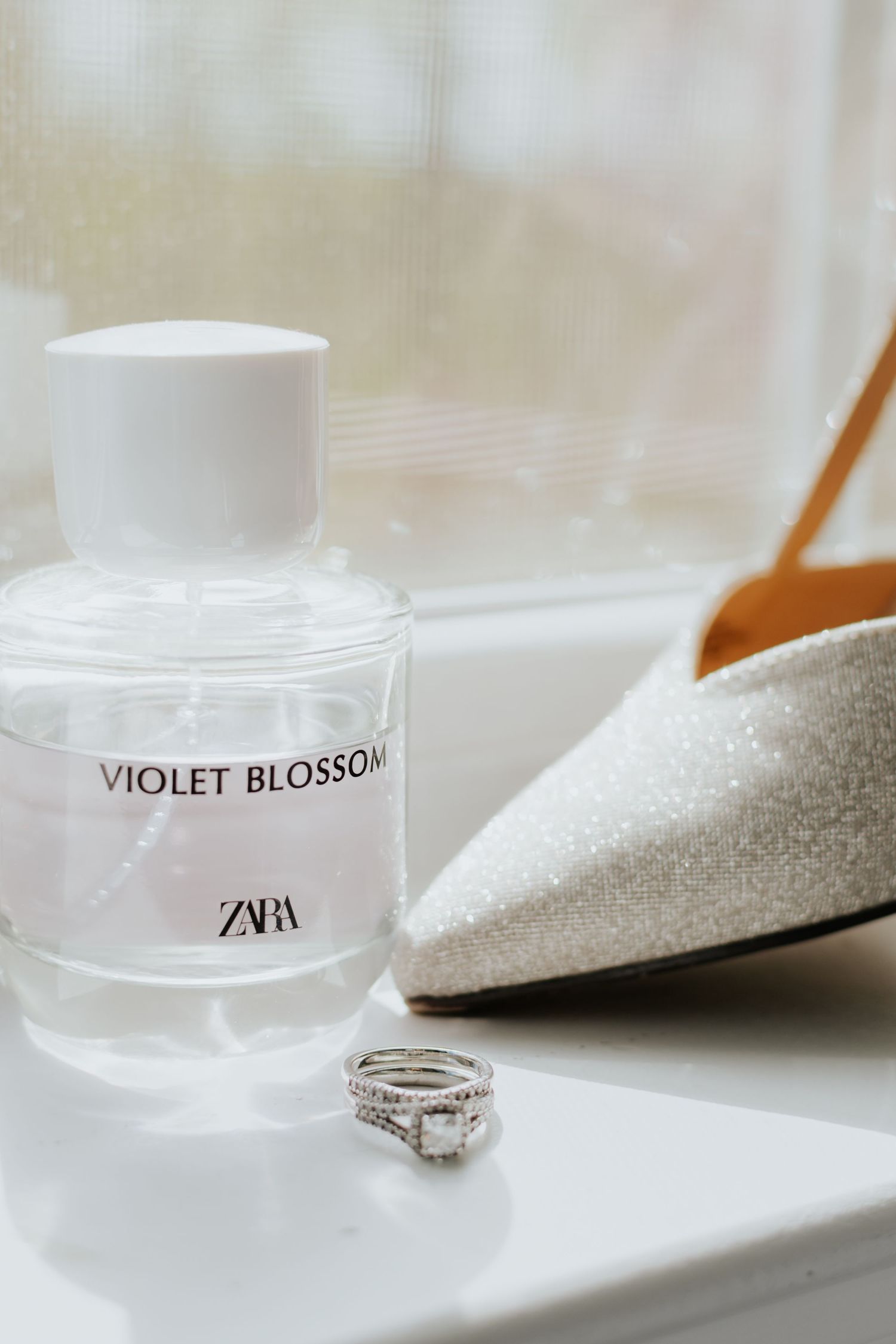 A Zara Violet Blossom perfume bottle sits elegantly beside a glittery shoe and diamond ring on a white surface.