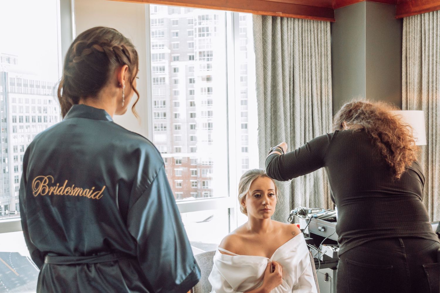 Makeup artist working on client's look in front of large windows in urban setting.