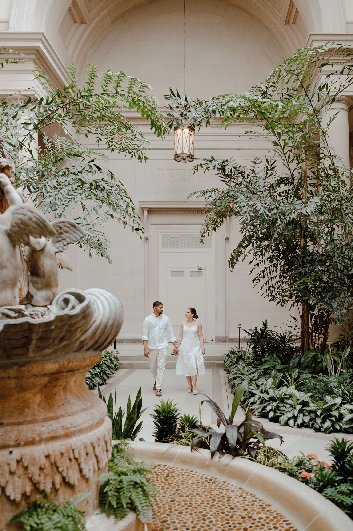 Couple in white stands in a lush indoor garden courtyard with a classical fountain and arched architecture.