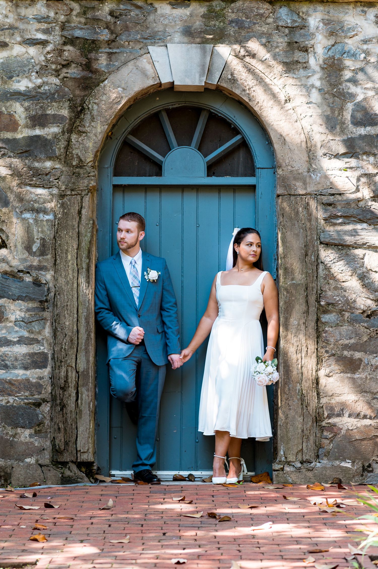 Wedding couple holds hands while standing in vintage blue doorway with stone arch.