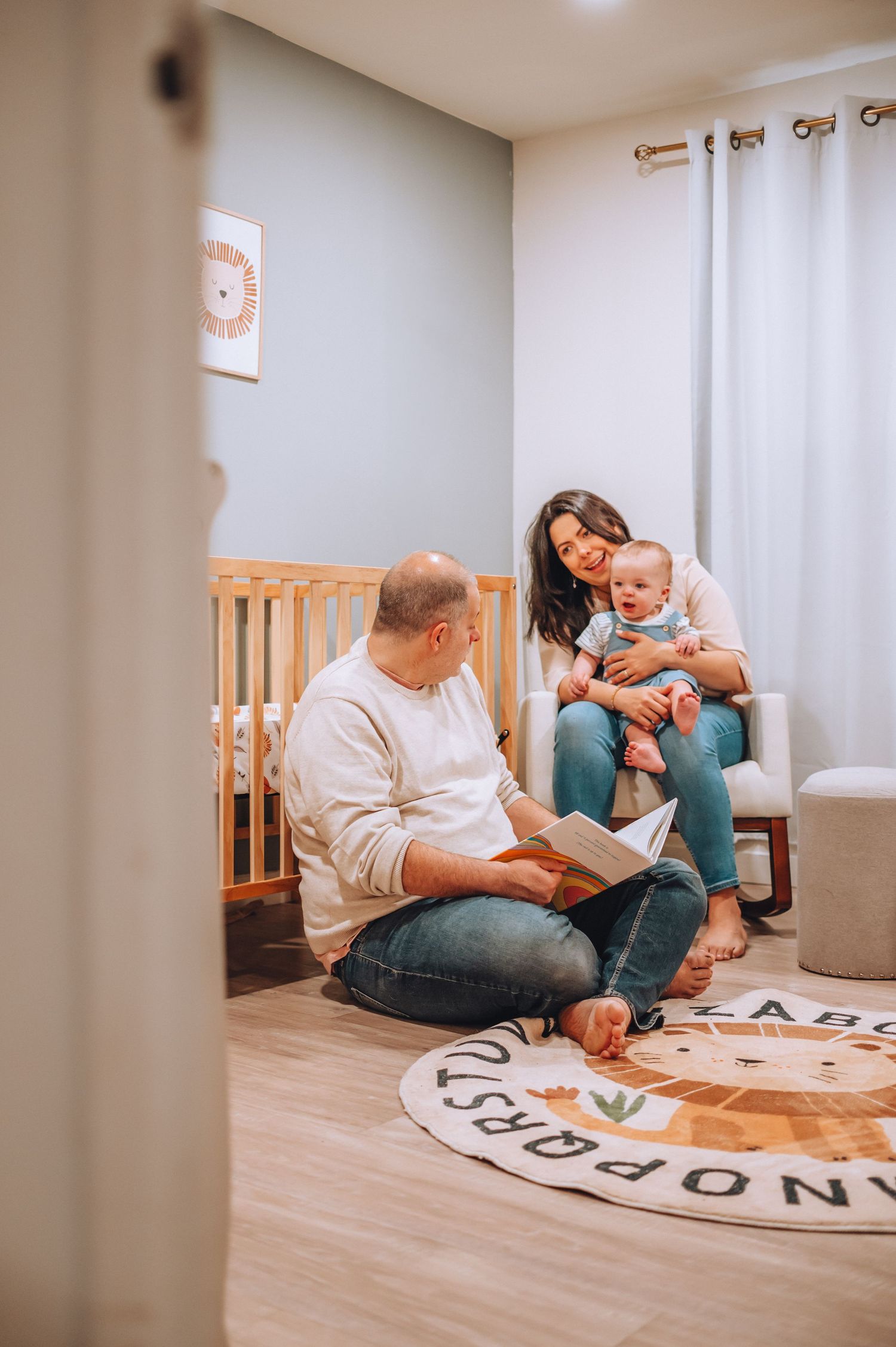 Candid family scene of story time in a modern nursery with white curtains and wooden floors.