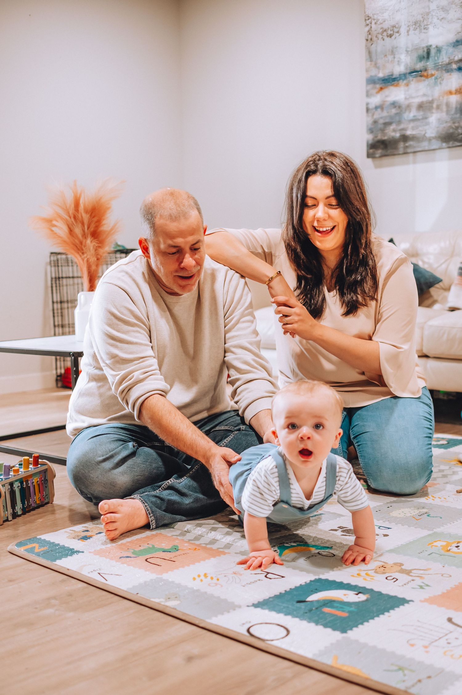 Baby crawls on colorful playmat while adults sit and play on hardwood floor in living room.