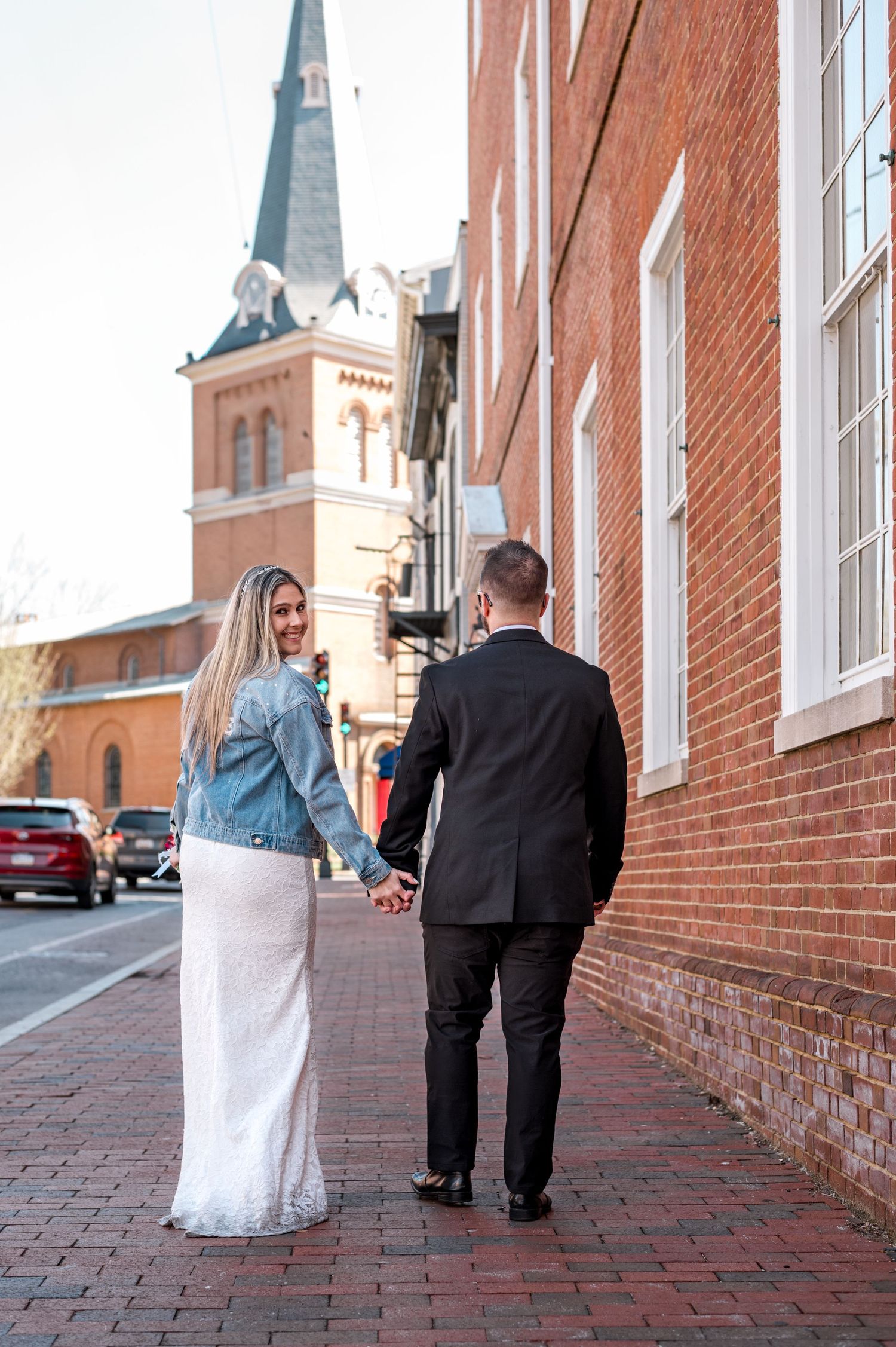 A couple walks hand in hand on a brick sidewalk with a historic church spire in the background.