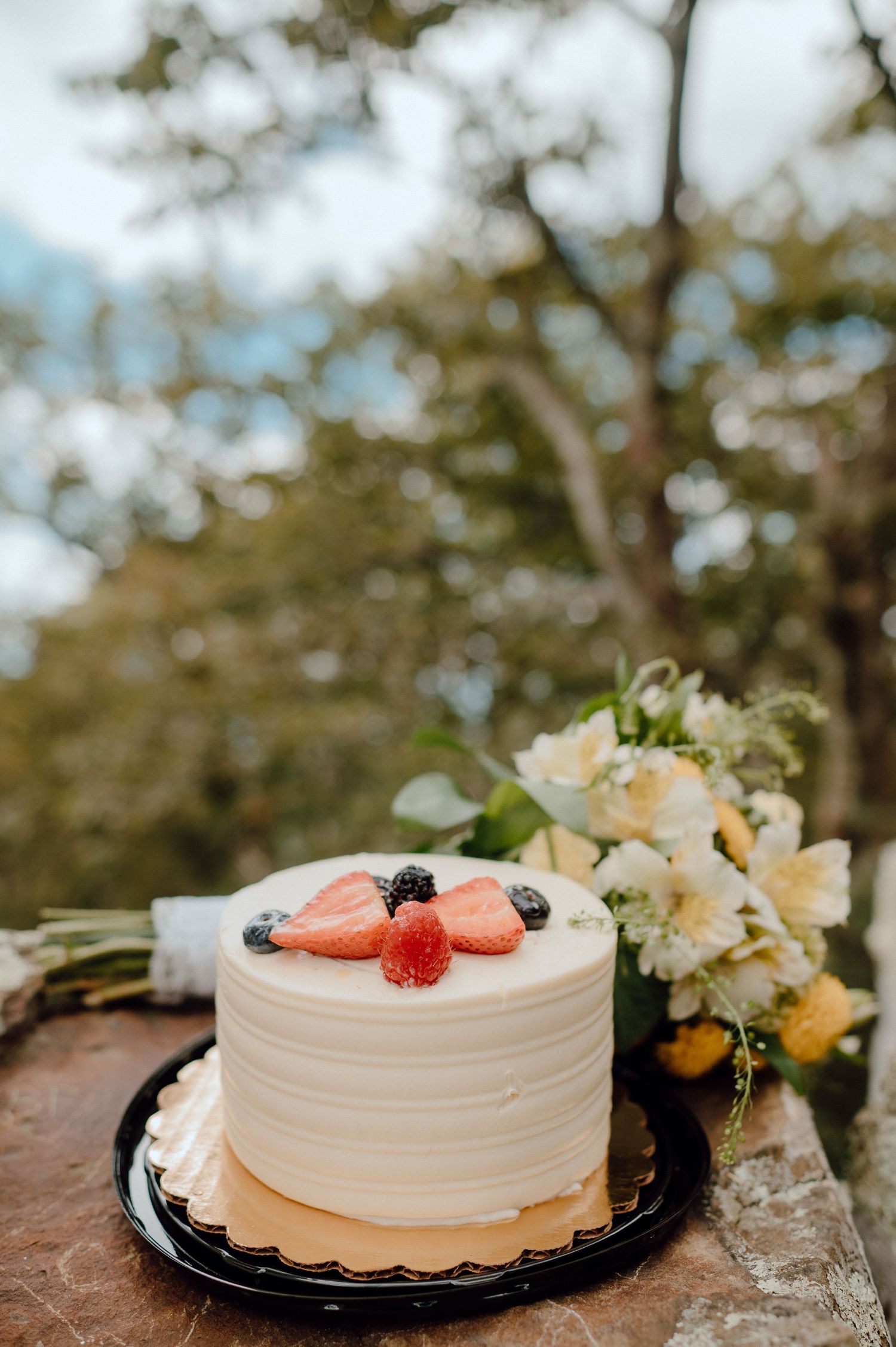 A small white frosted cake decorated with fresh strawberries and blackberries sits beside floral arrangements outdoors.