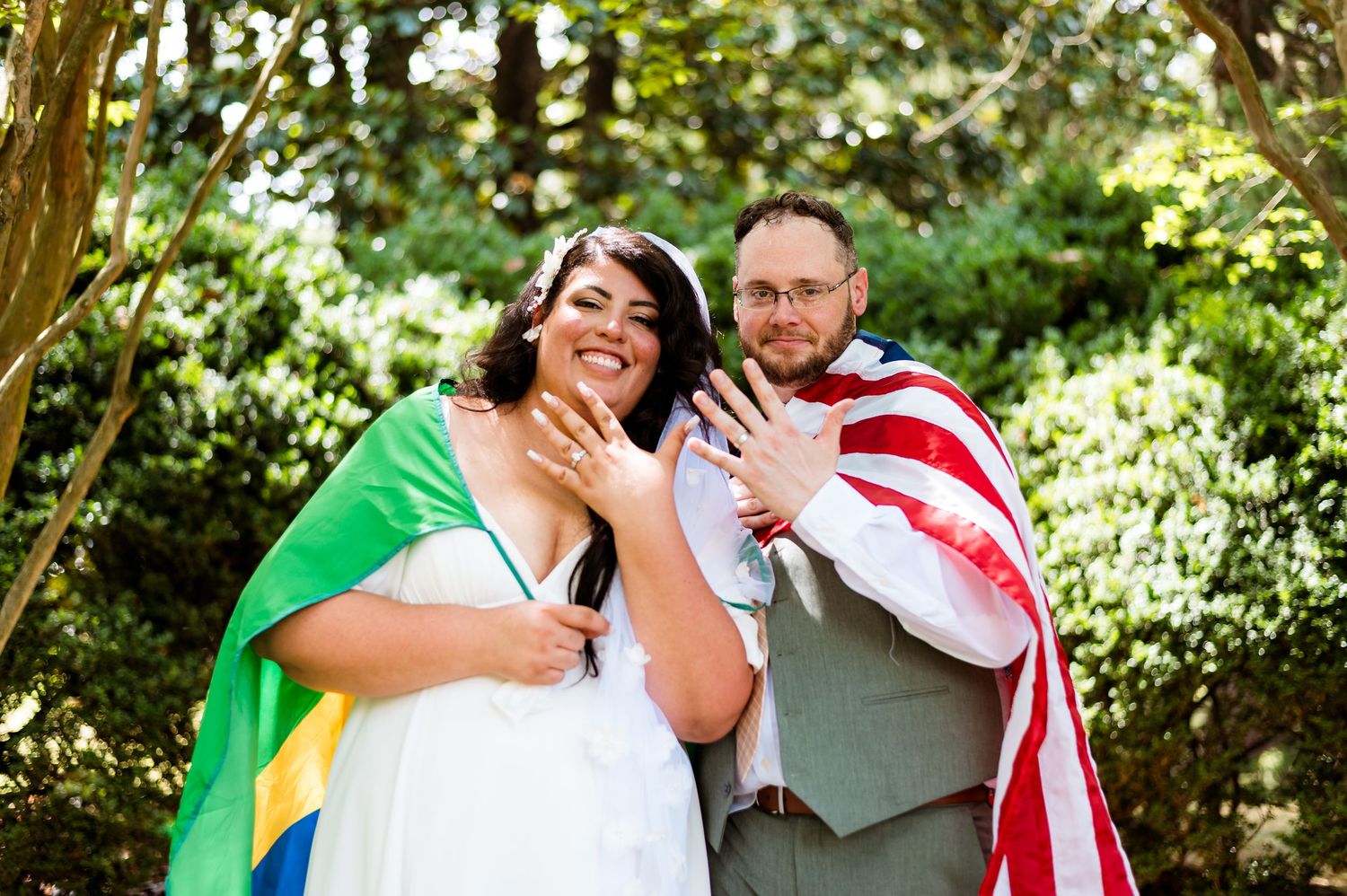 Two people draped in American and Brazilian flags stand together outdoors in a woodland setting.