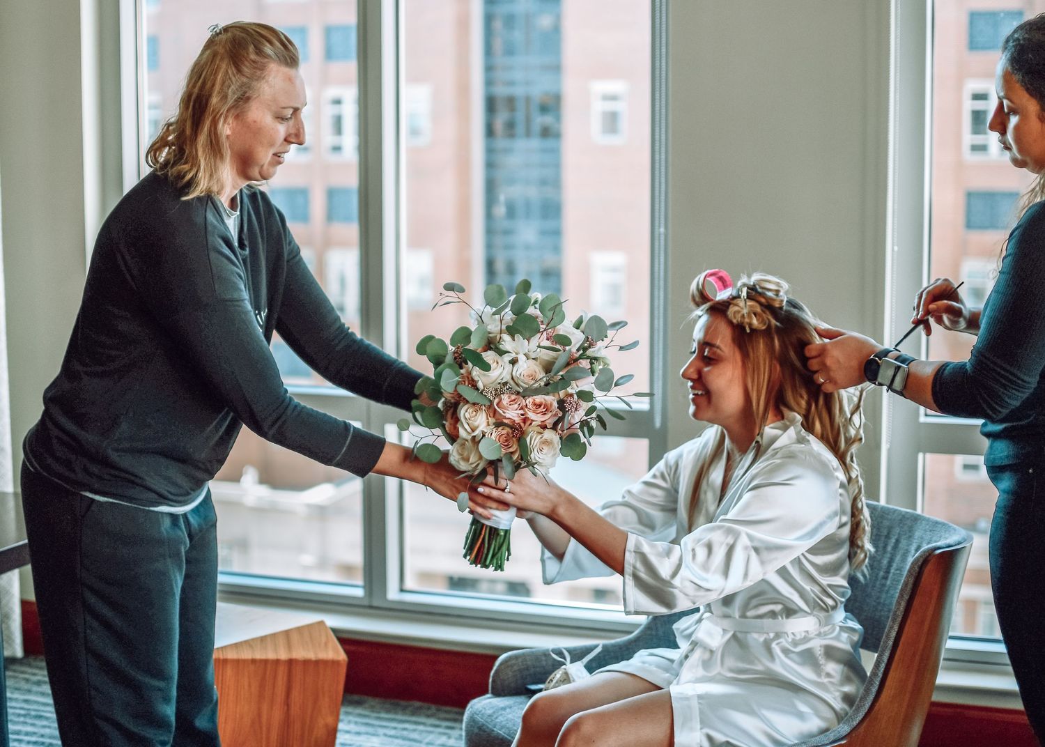 A wedding preparation scene shows a floral bouquet being handed over while hairstyling is done in a bright room.