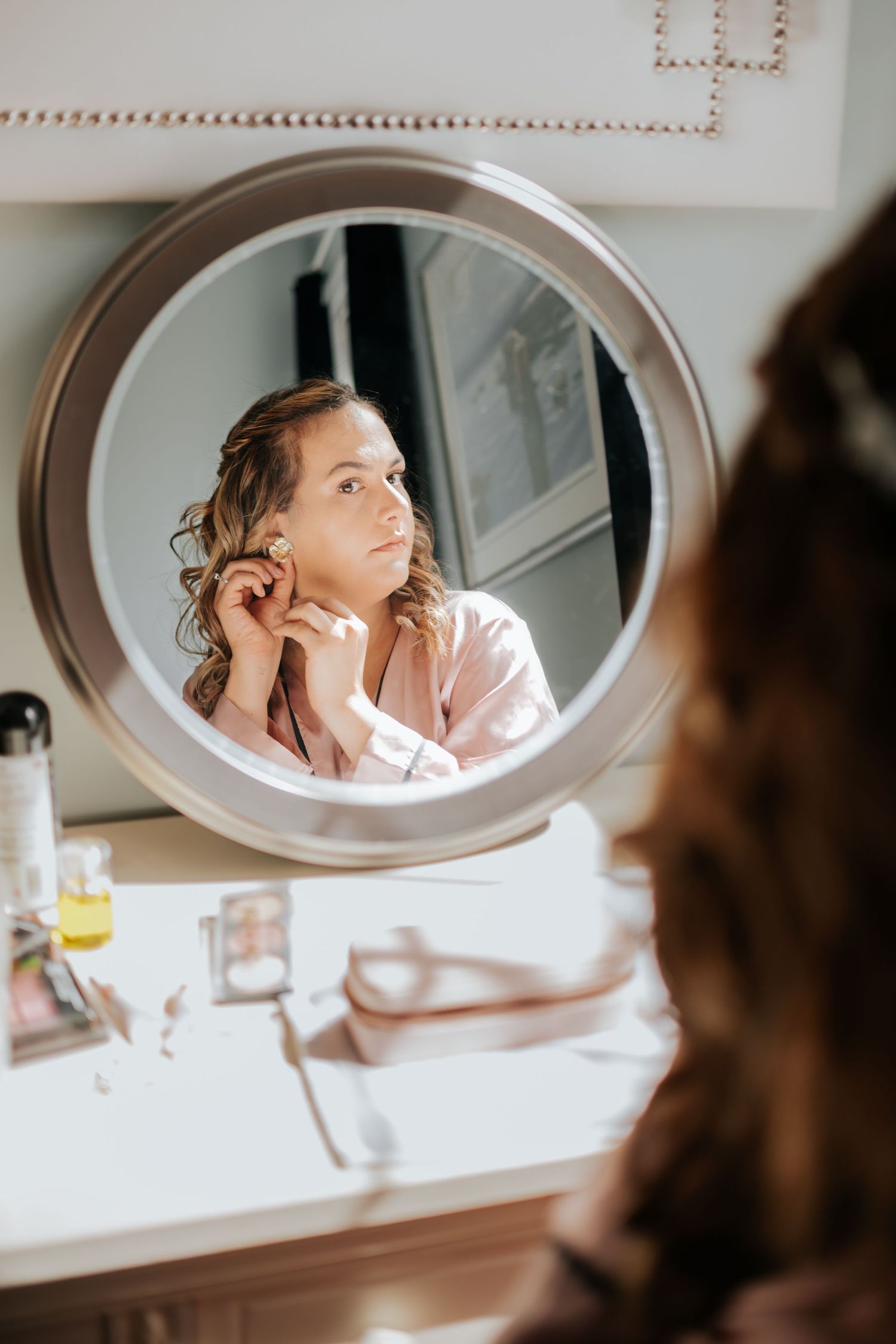 Reflection in round mirror shows person in pink robe sitting at vanity table with beauty products.