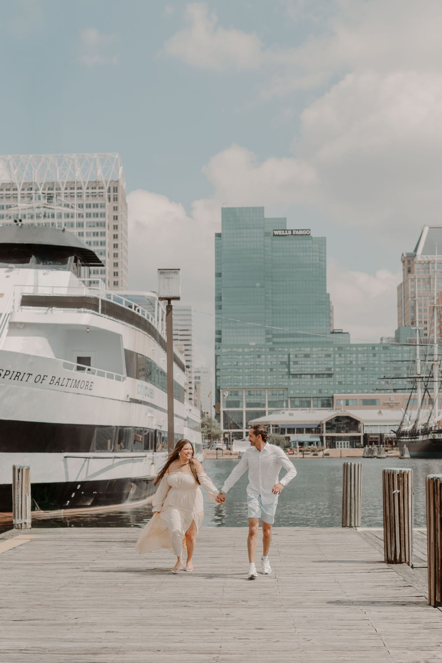 A couple runs playfully along a waterfront boardwalk with the Spirit of Baltimore cruise ship and city skyline visible.