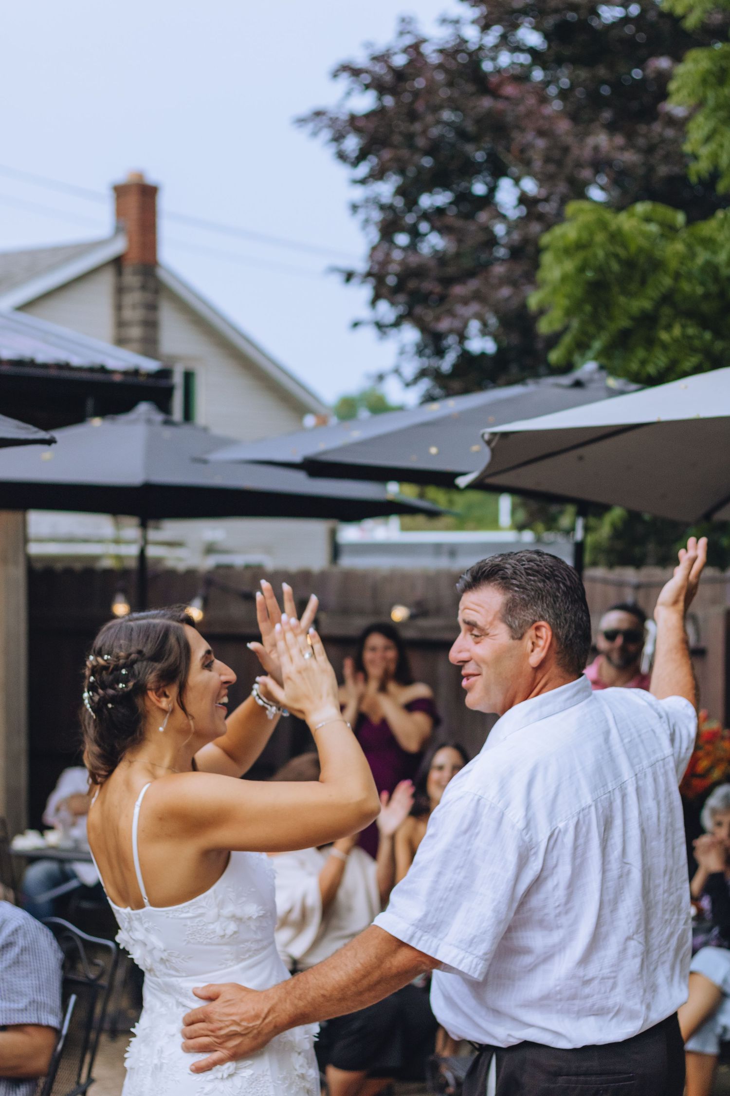 A wedding celebration at an outdoor venue with guests dancing under patio umbrellas and trees.