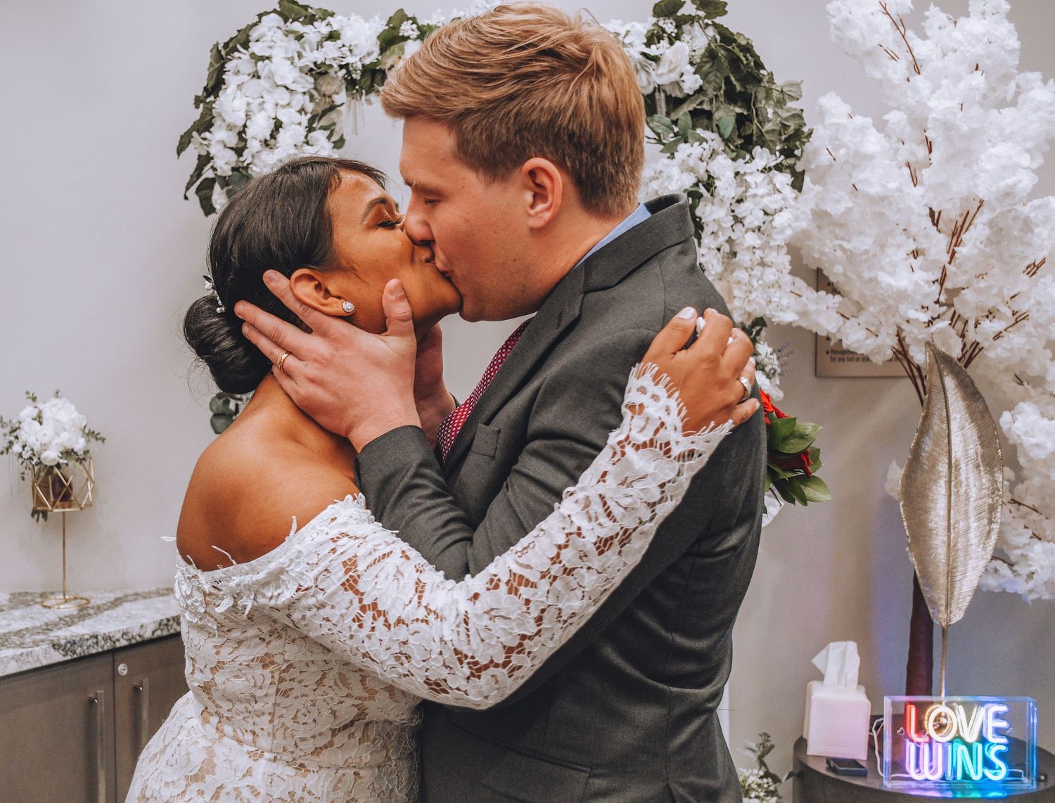 Newlyweds share their first kiss under a decorative arch with white flowers.