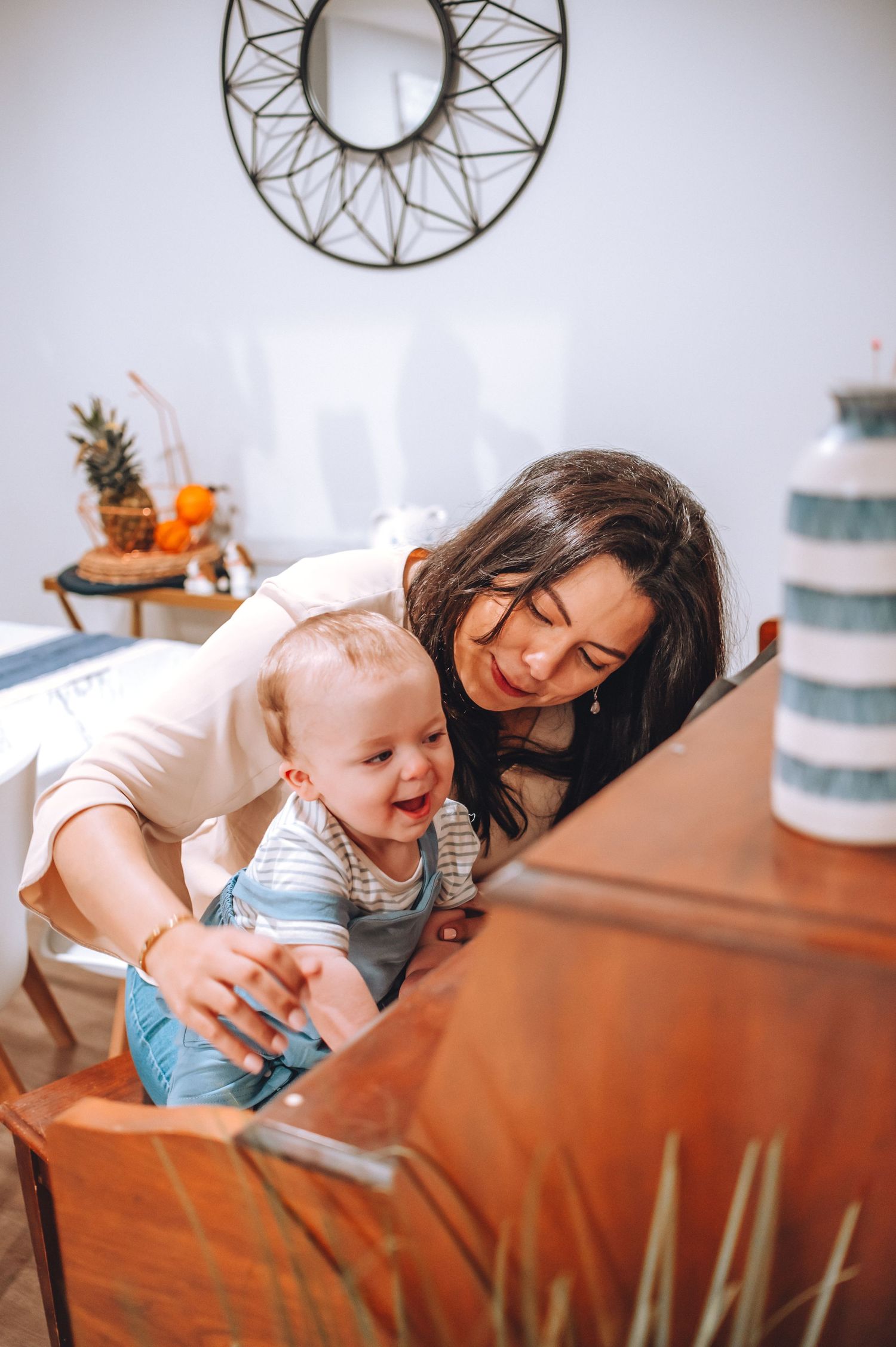 Parent and baby playing together at wooden piano in bright room with decorative mirror.