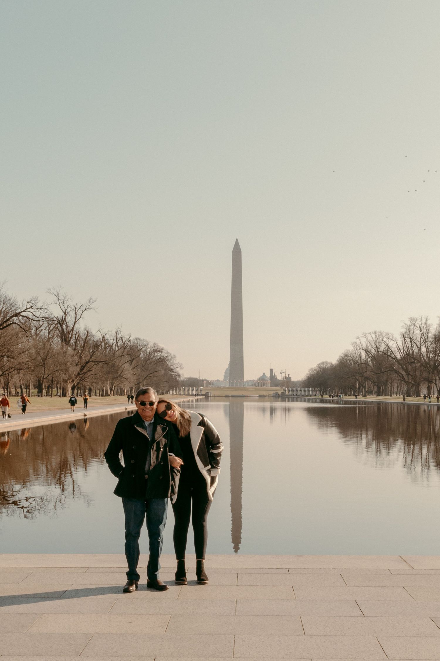 Couple stands near reflecting pool with Washington Monument in background on misty morning.