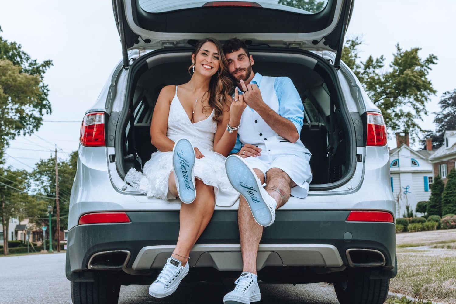 A couple wearing white outfits sits in the open trunk of an SUV while eating snacks together on a sunny day.