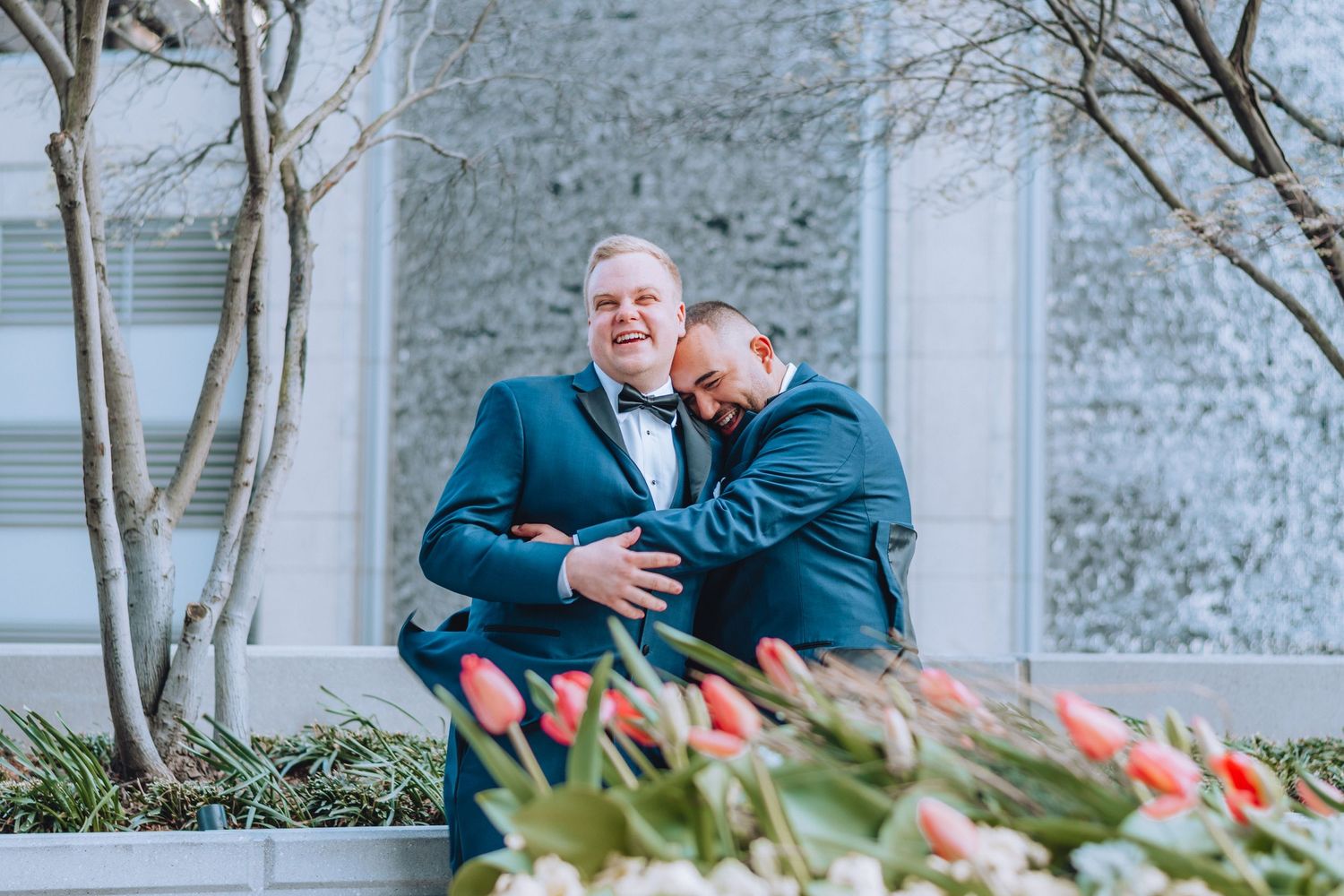 Two people in formal attire share an intimate moment outdoors near white stone walls and red tulips.