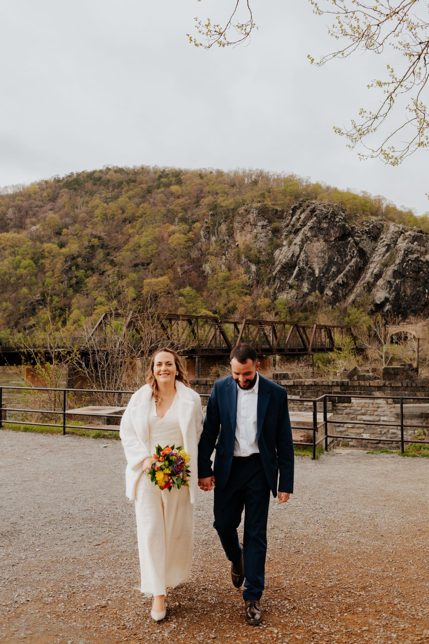 A couple walks hand in hand on a gravel path with fall foliage and a rustic bridge in the background.