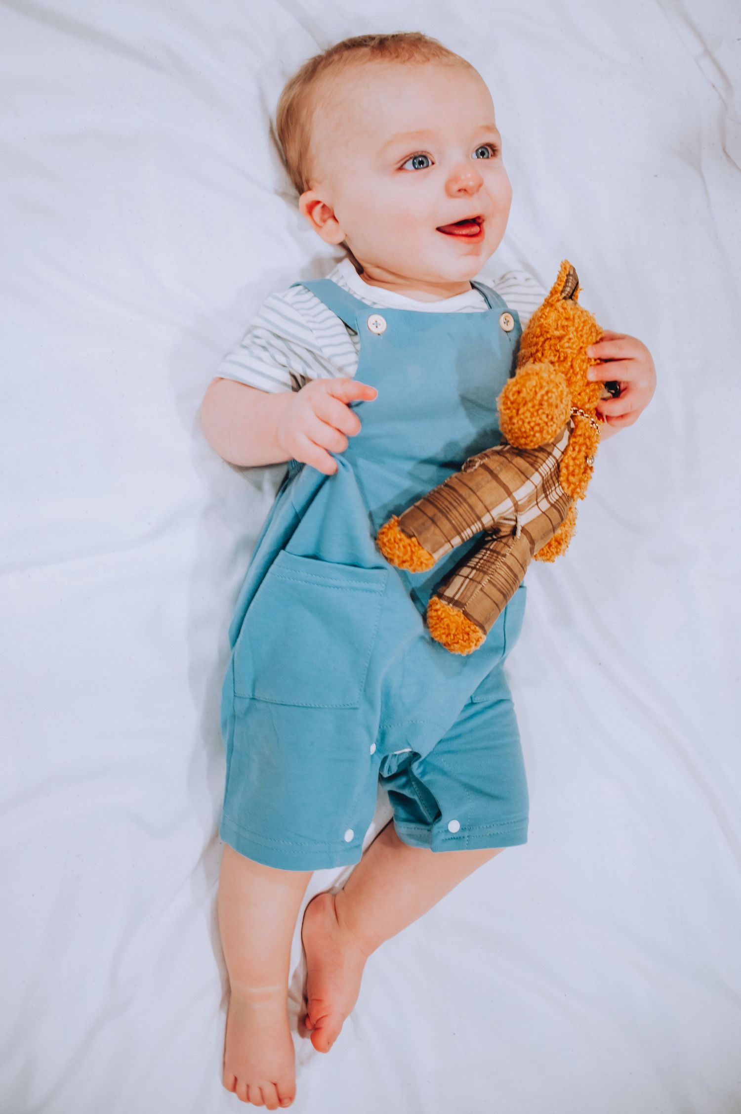 Baby in blue overalls holds orange plush toy while sitting on white bedding.
