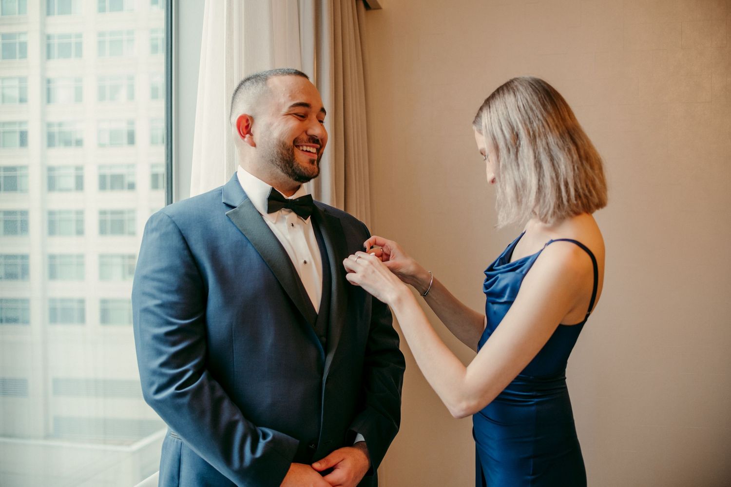 Two people in formal attire share a happy moment by a sunlit window.