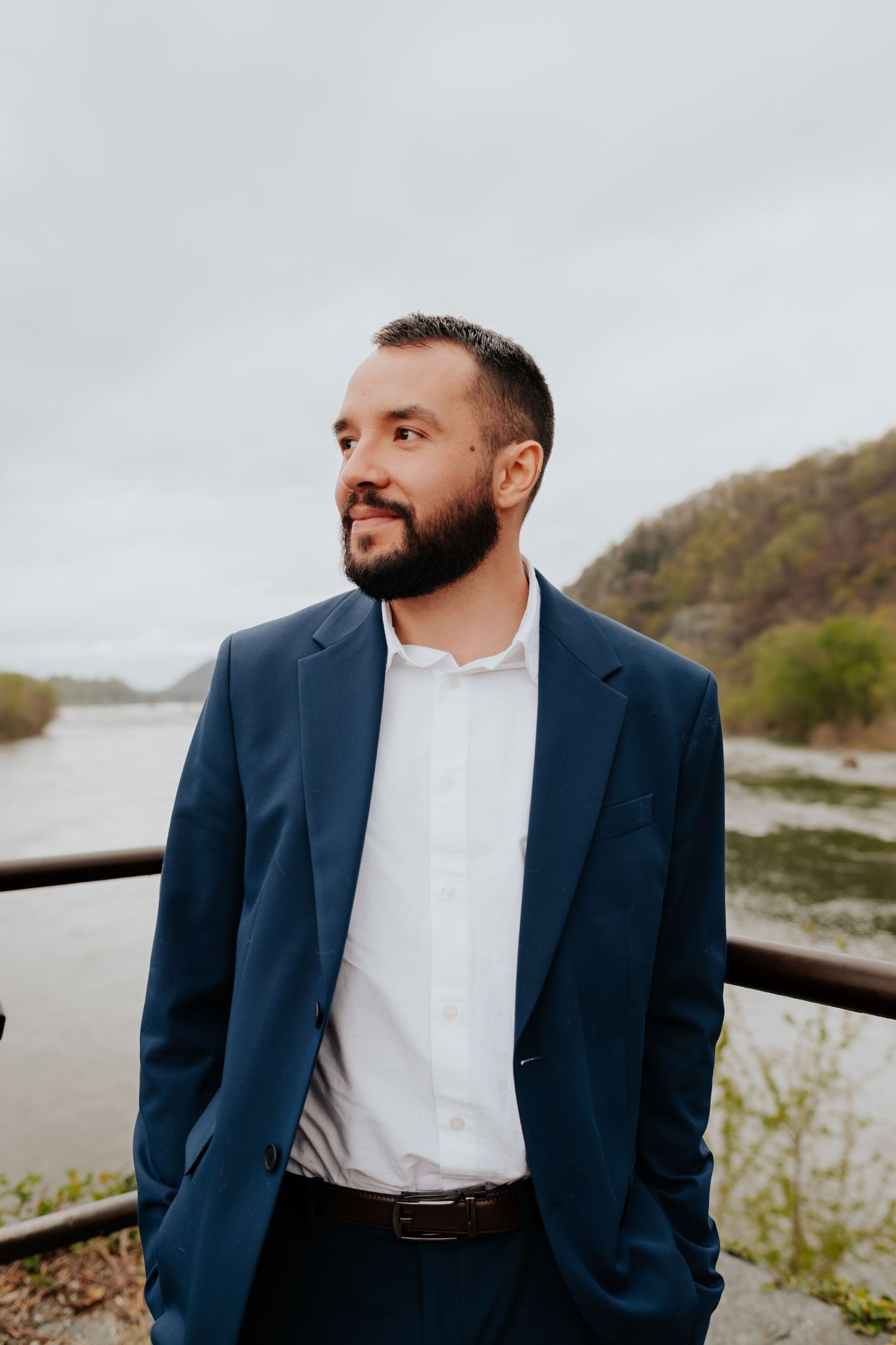 A person in a navy blue blazer and white shirt looks contemplatively into the distance near a river on an overcast day.