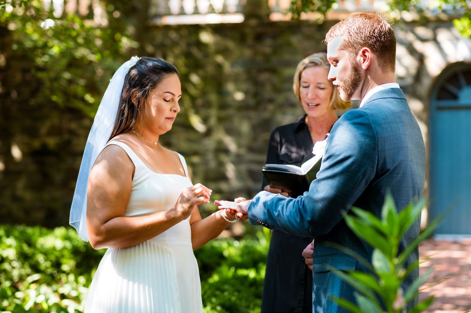 Wedding ceremony taking place in a garden with exchange of rings in front of stone architecture.