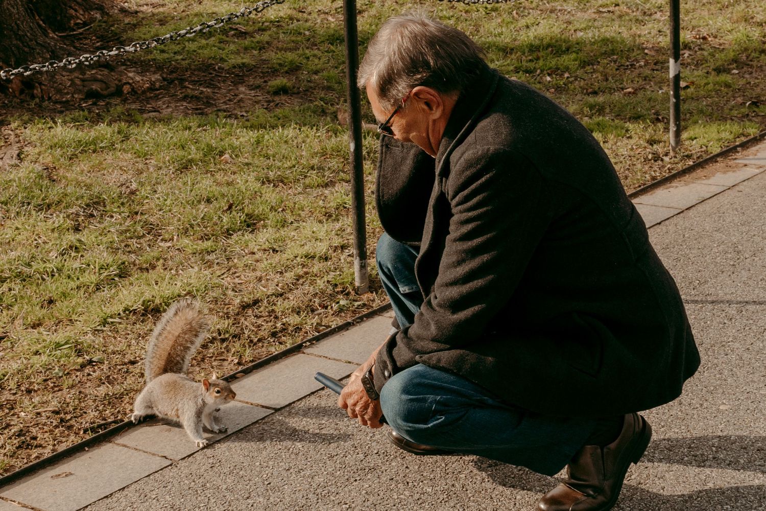 Person in dark coat crouches down to feed a small squirrel on a walkway.